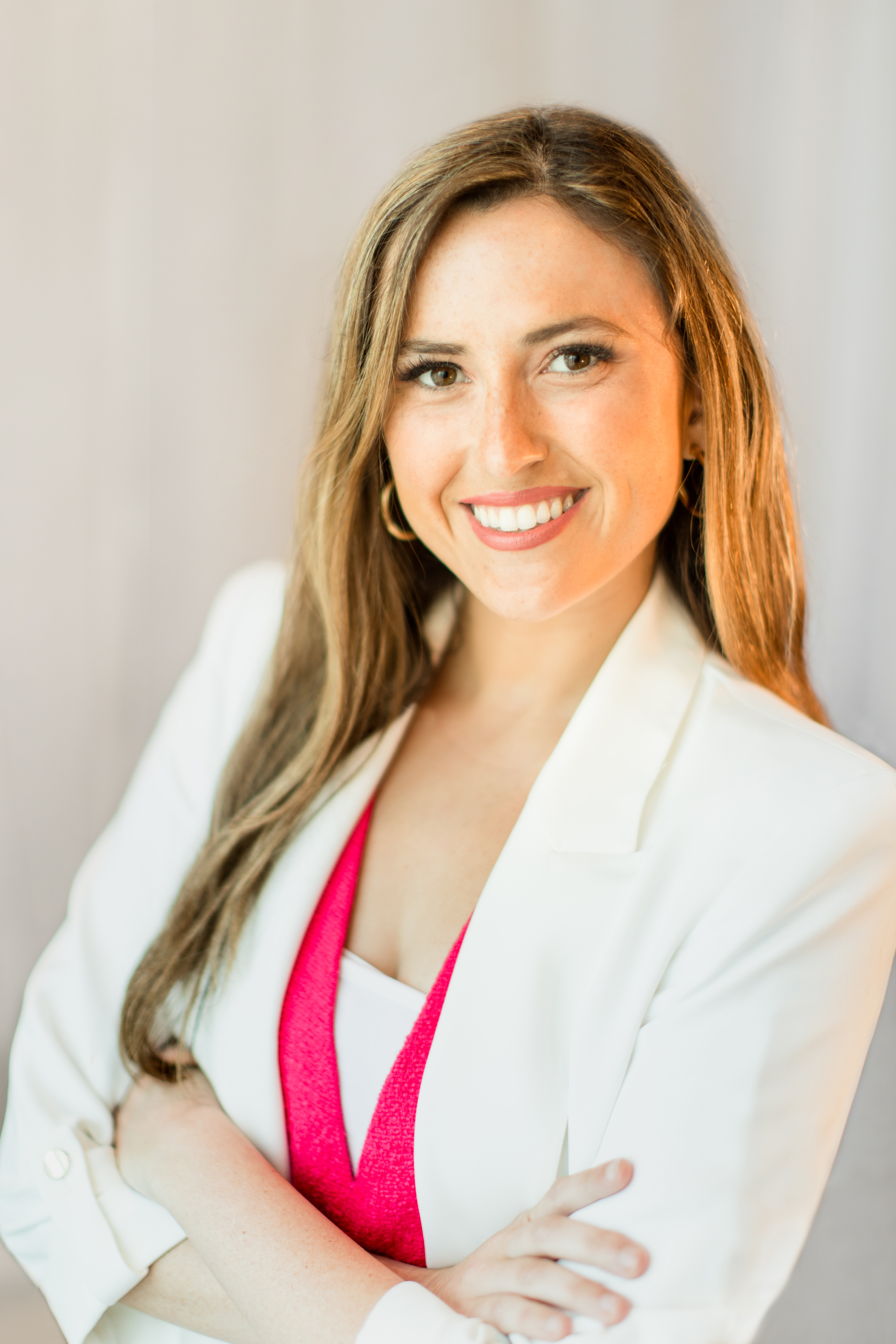 Katherine is wearing a white coat with a pink top and gold earrings. She is smiling with her arms crossed.