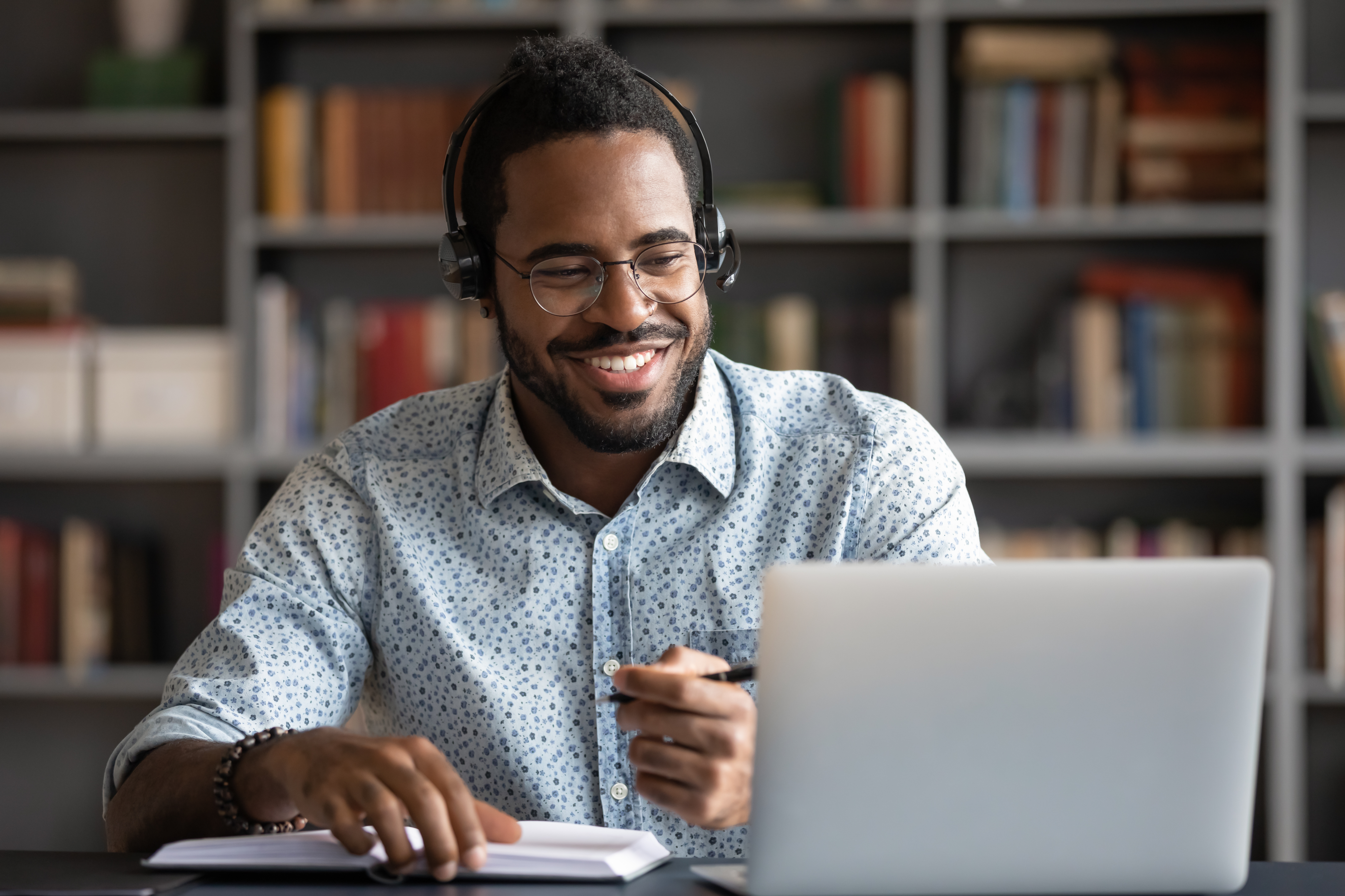 Image of a Black, professional man wearing headphones and smiling at a laptop while making notes with a pen