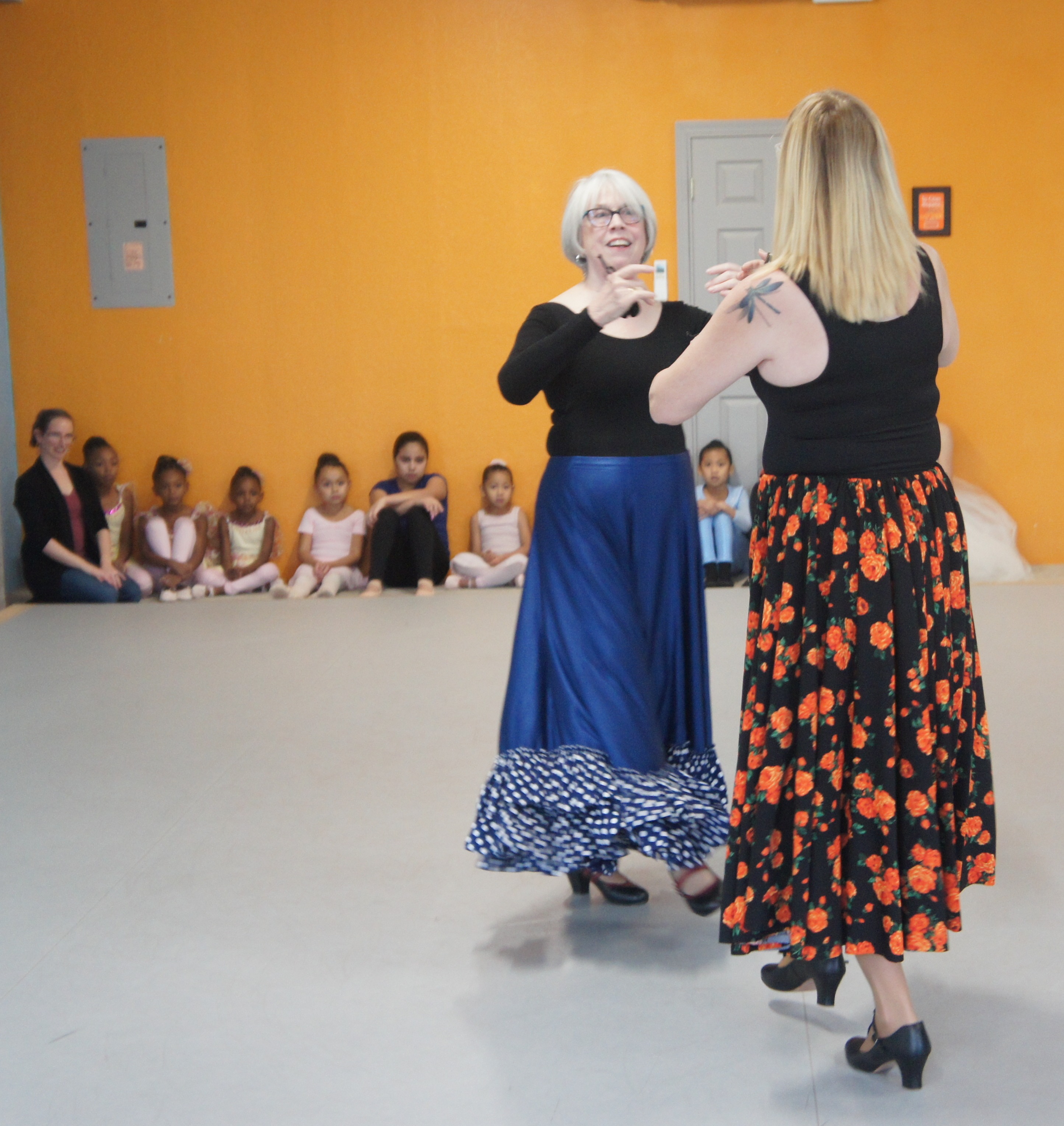 At a Find Your Center studio performance in 2019, two adult women dance together in flamenco skirts and shoes on a light gray floor, while multiracial children in yellow, pink, black, and blue ballet clothes sit with their instructor by an orange wall and watch