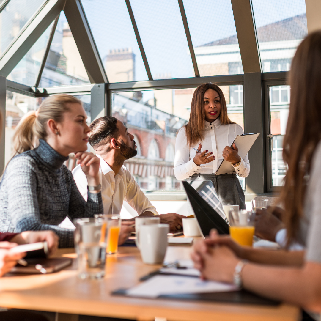 woman stands at a table talking, whilst adults sitting at the table listen