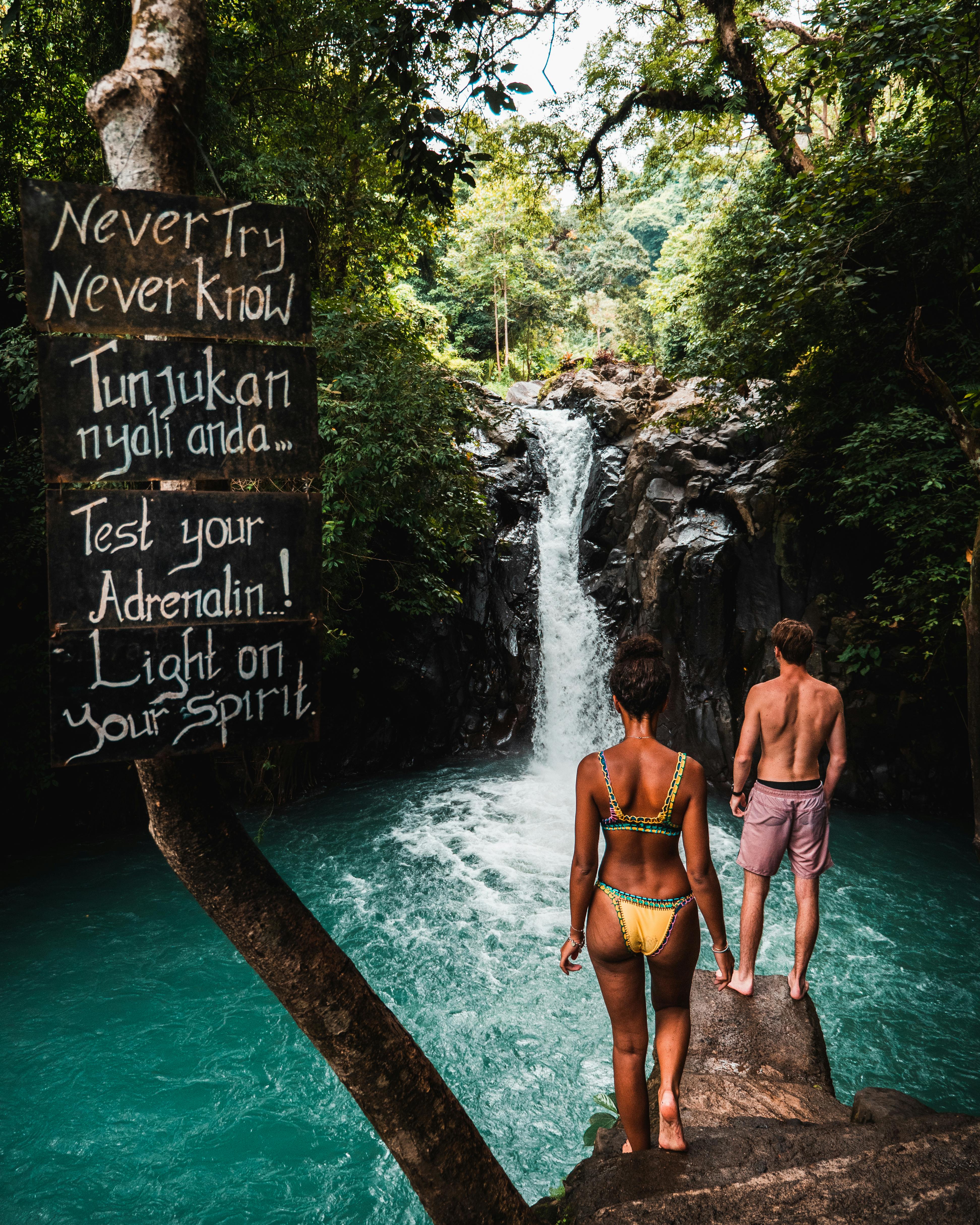 man and woman stand at bali waterfall edge considering to jump, there is a sign that says never try never know