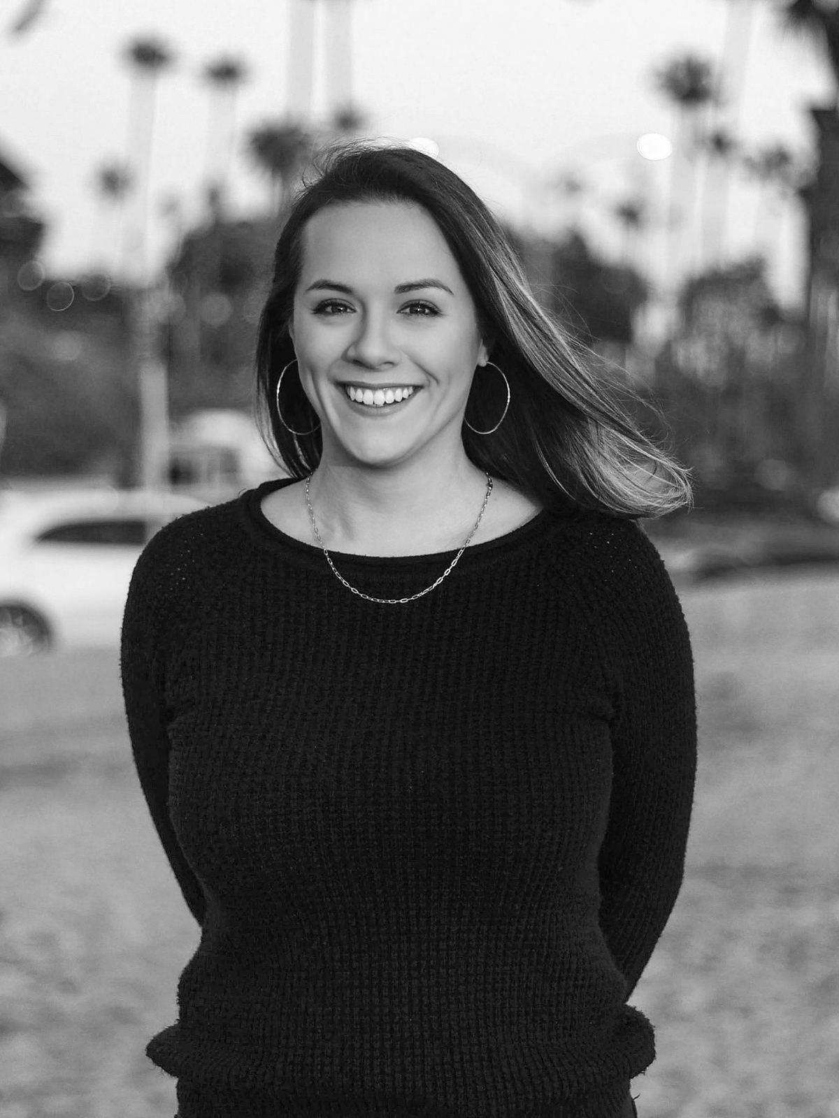 Woman wearing a black sweater and gold necklace smiling at the beach in front of palm trees