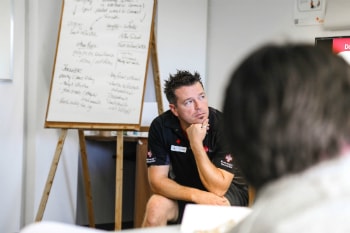 A Canadian Red Cross instructor wearing a black t-shirt with a Red Cross symbol is actively listening to a student. In the background, there is a board with notes from a classroom. 