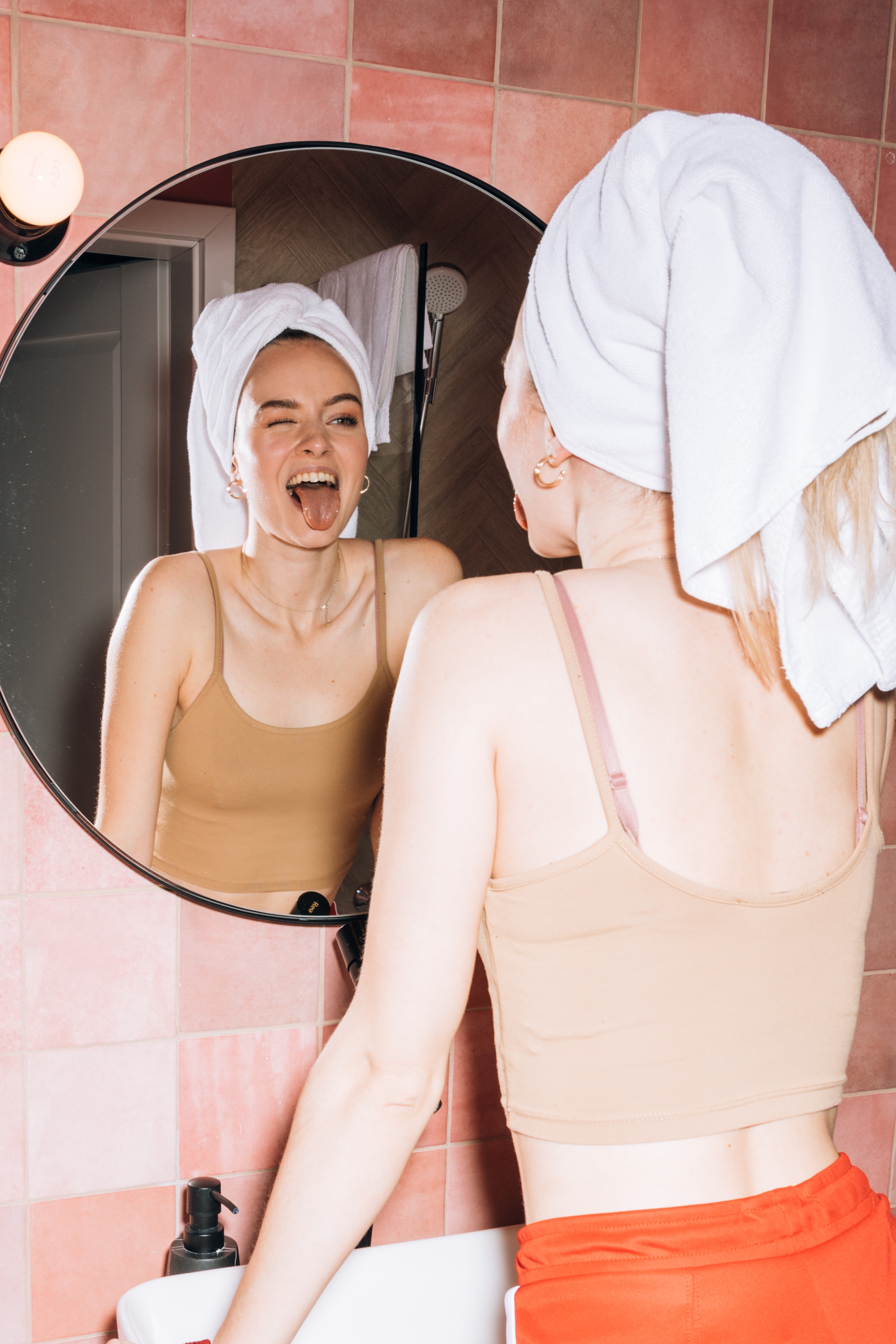 Girl with her head wrapped in a towel is looking in the bathroom mirror and sticking her tongue out and smiling.