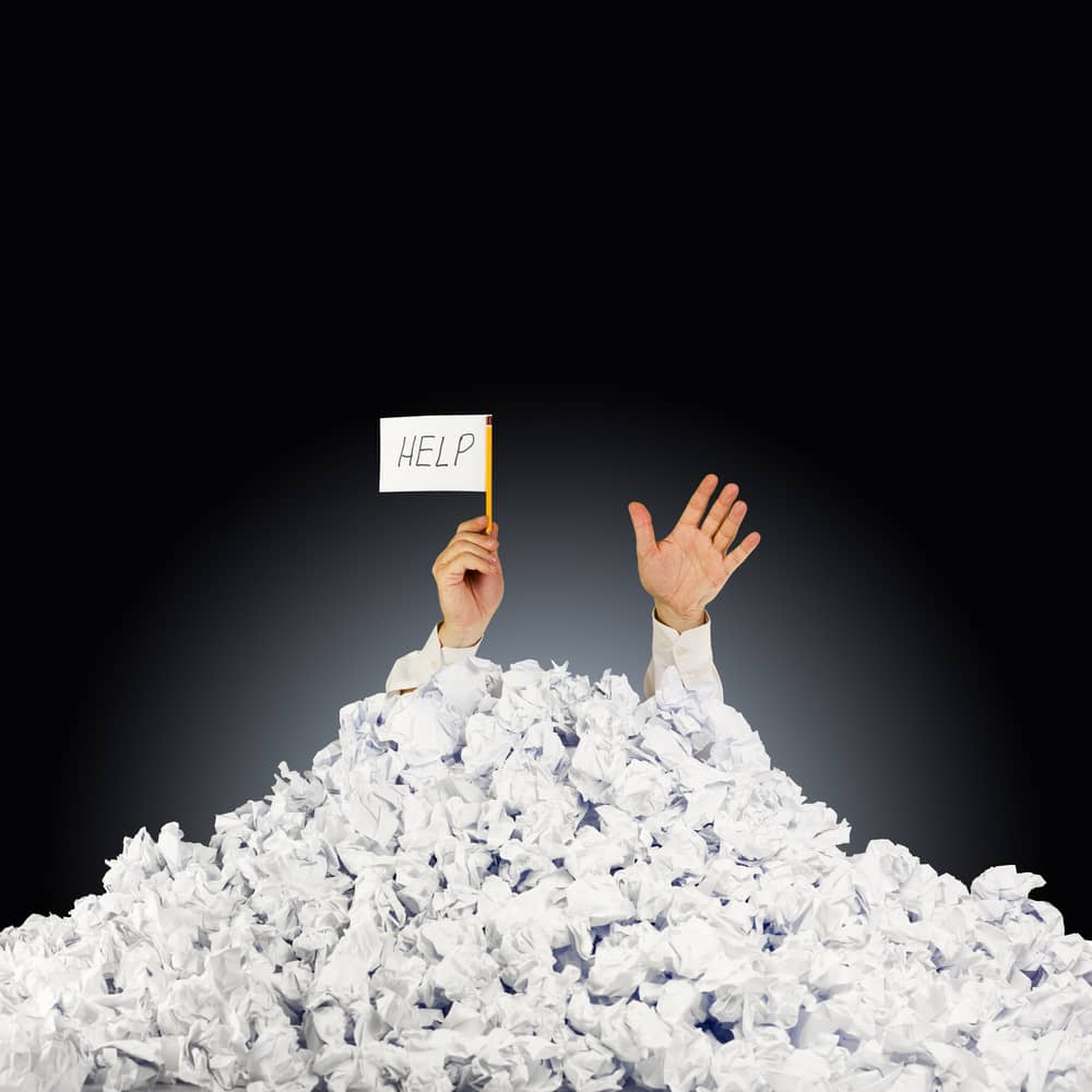 man buried in pile of crumpled papers. Only his hands are sticking out of the top of the pile. He is holding a tiny white flag that says 