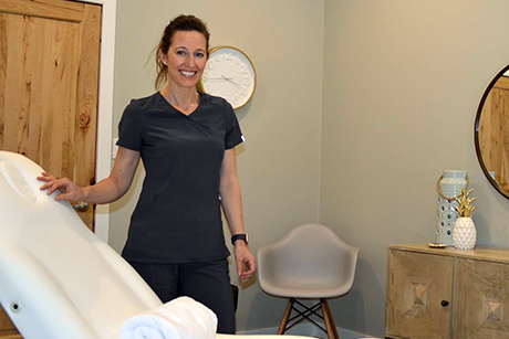 Brenda stands in her medical aesthetics clinic, smiling and wearing dark scrubs.