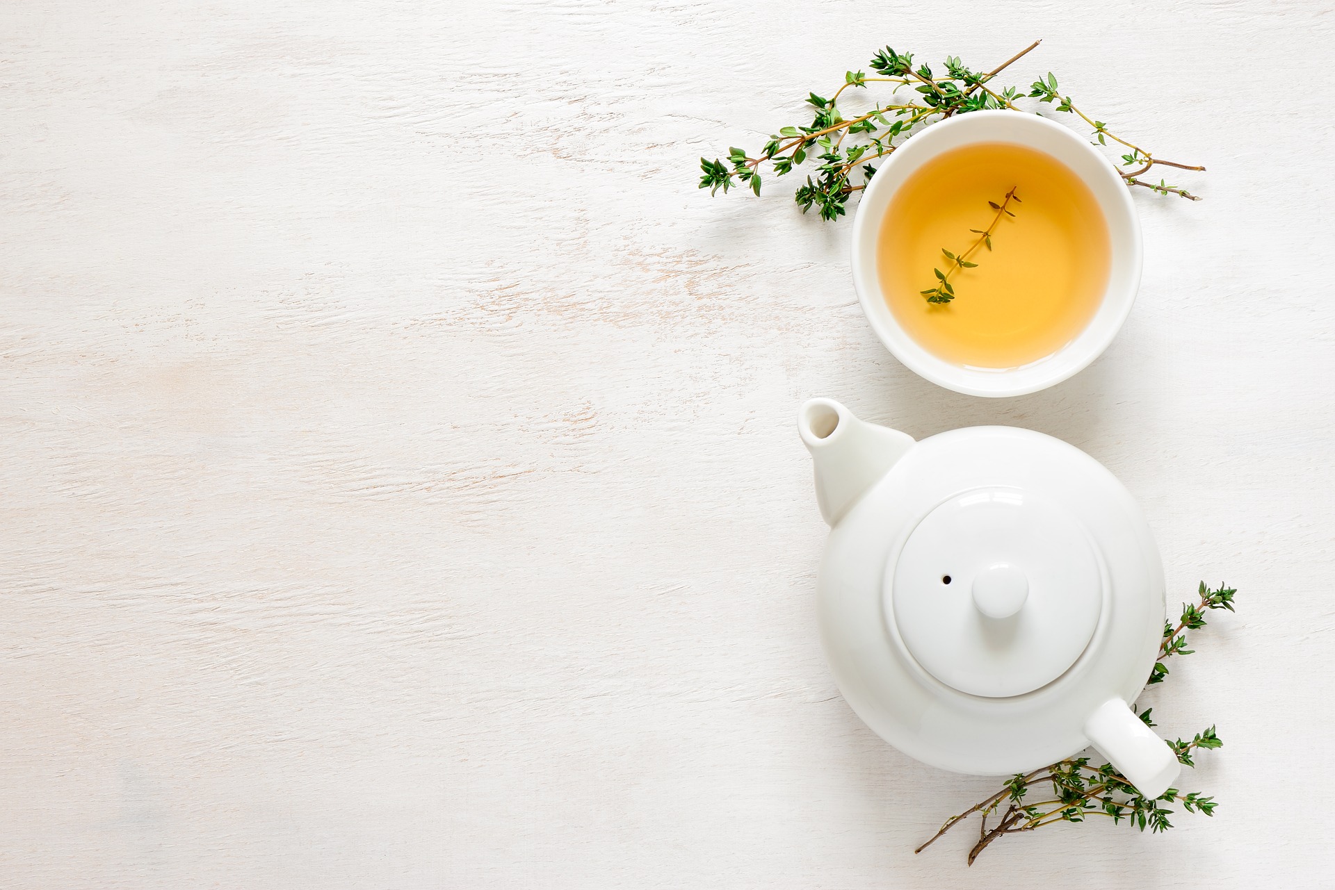 Looking down on a neutral colored background with a white teapot and cup of tea with sprigs of thyme around them