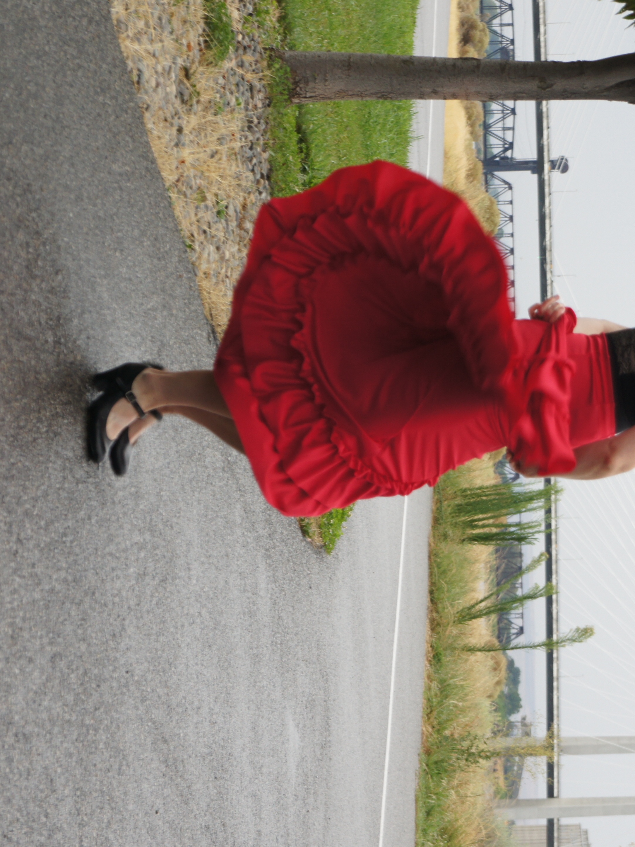 Instructor Kate Feinberg Robins wooshes a red flamenco skirt in a circular motion while performing footwork in black flamenco shoes on a bicycle path in front of a bridge
