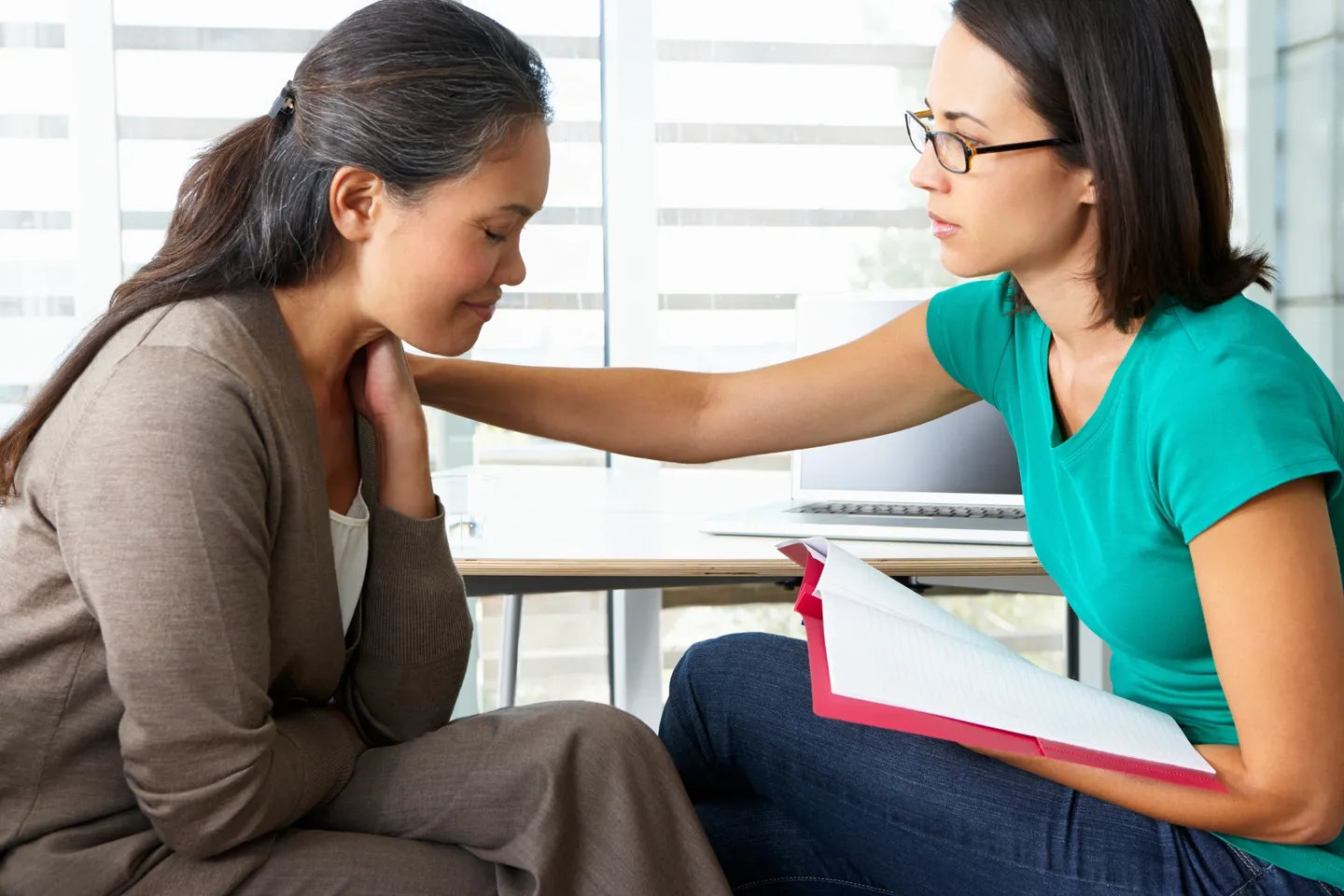 two women sitting in a room, one woman appears upset, the other has a folder open and is reaching out to comfort the first woman