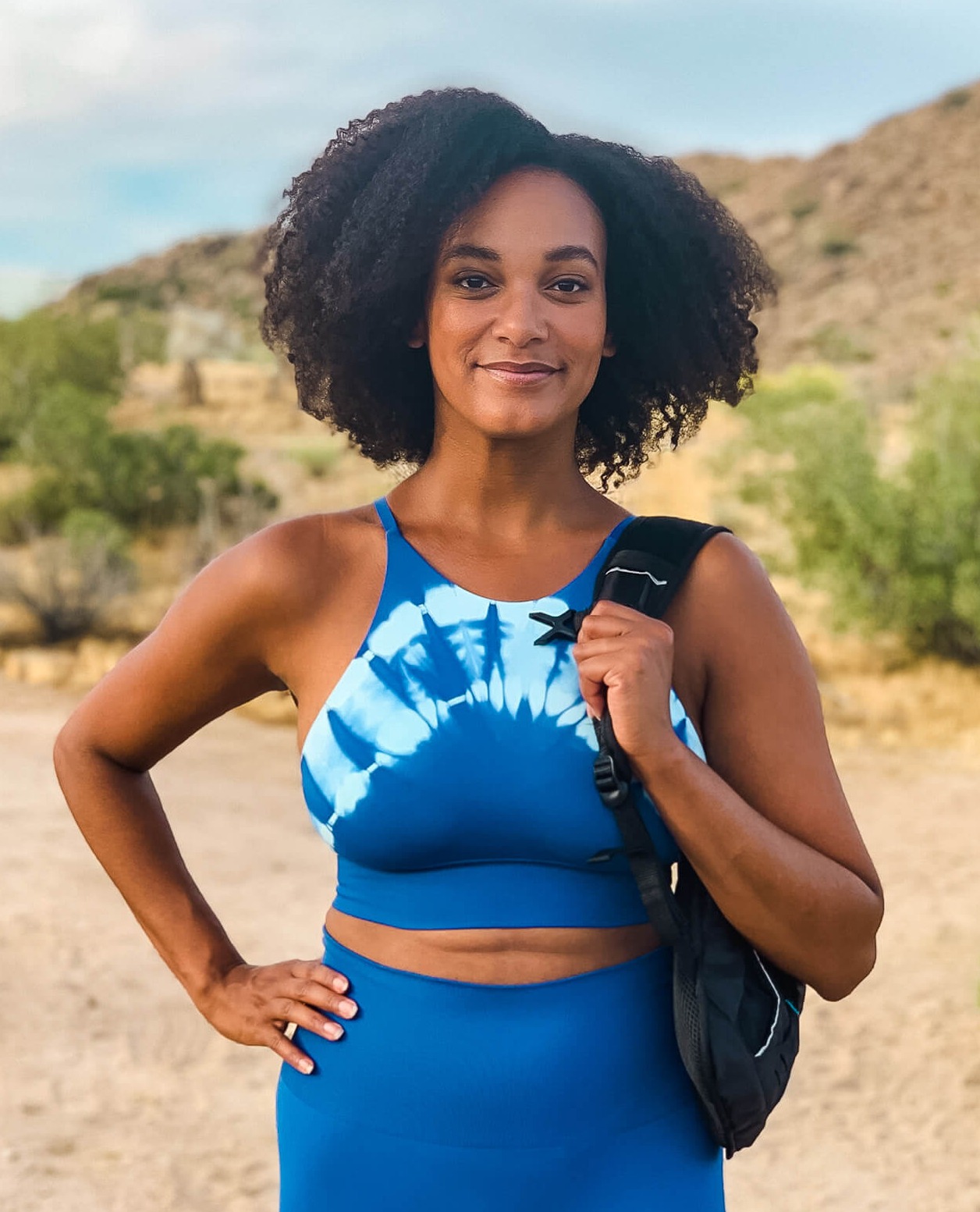 Black Hiker getting outdoors in Joshua Tree National Park holding a backpack, hand on her hip, wearing an afro