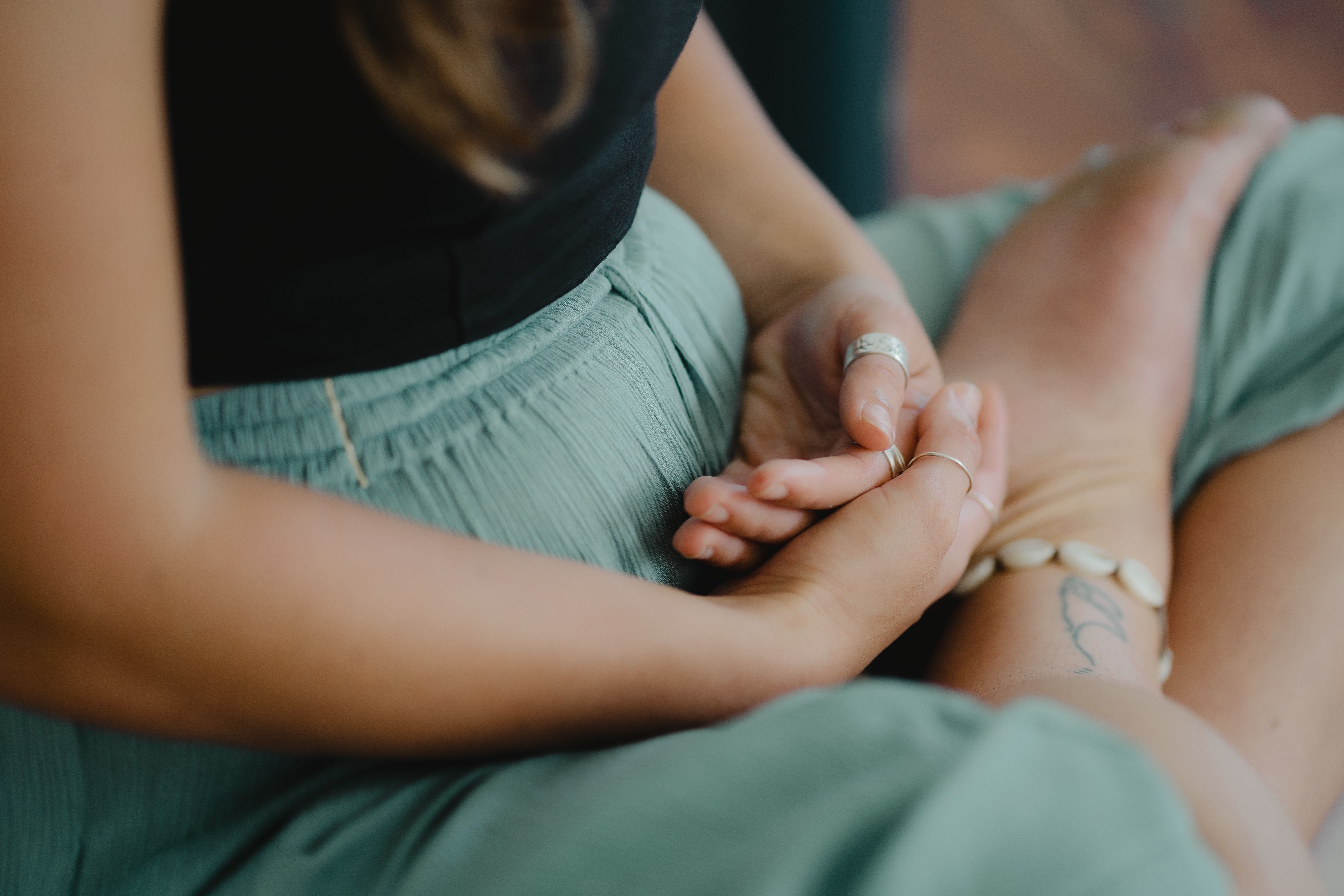 Woman is sat cross legged in a meditating pose. She is practicing mindfulness. She wears a shell anklet. She suffers from anxiety is using mindfulness to help her. Her palms are in her lap. She wears a black top and green trousers. She seems peaceful. 