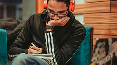 A man writing in a notebook in a cafe while listening to an online course using headphones.