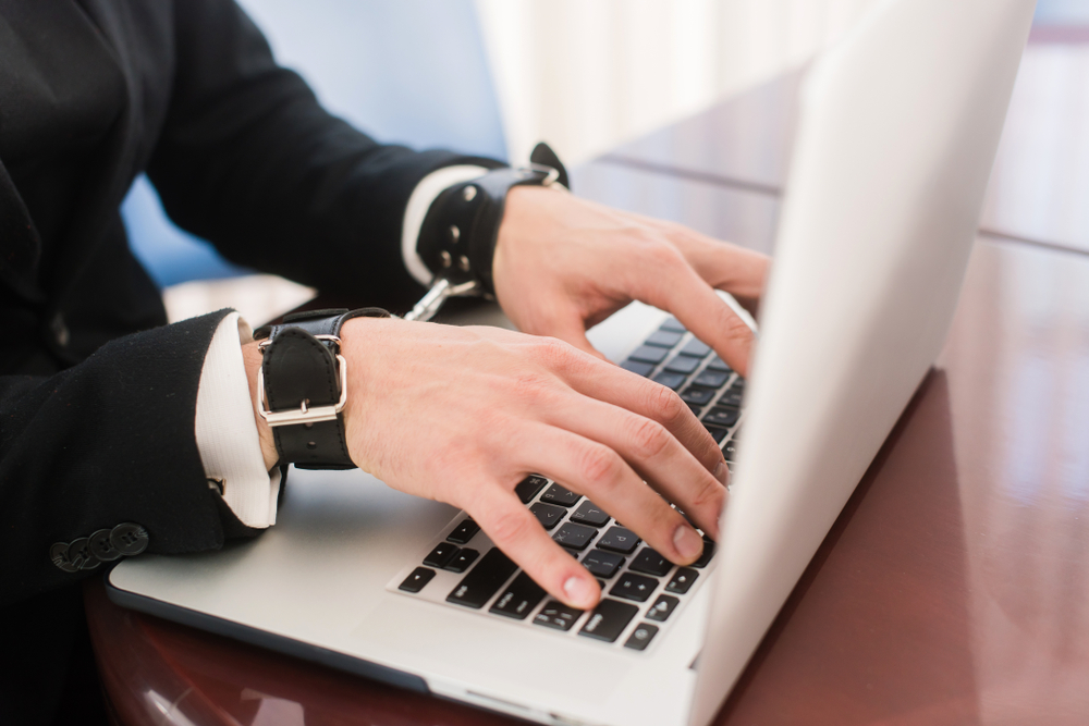 Person in suit wearing black wrist restraints while typing on a computer