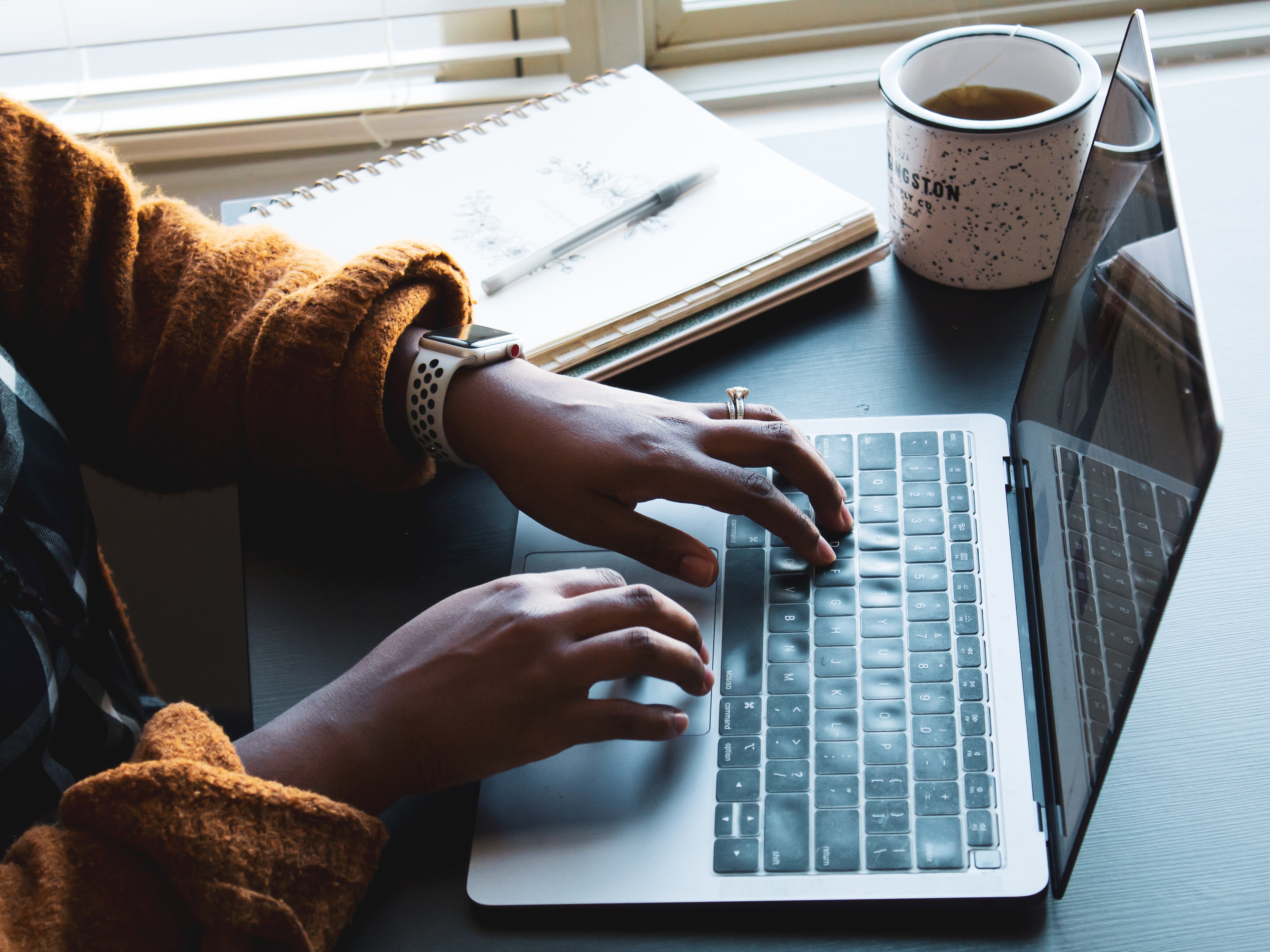 Person typing on a computer screen with notebook and tea. Daniel Thomas on Unsplash.