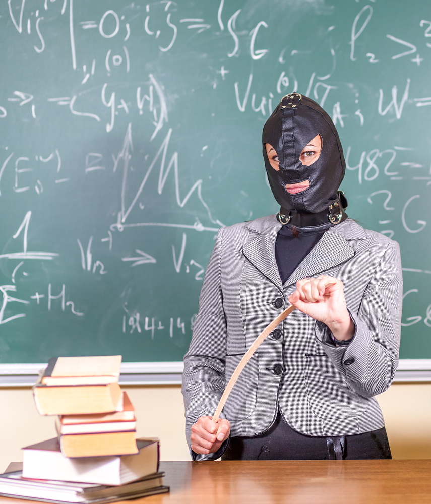 A teacher wearing a collar and gimp mask stands in front of a chalkboard full of equations. 