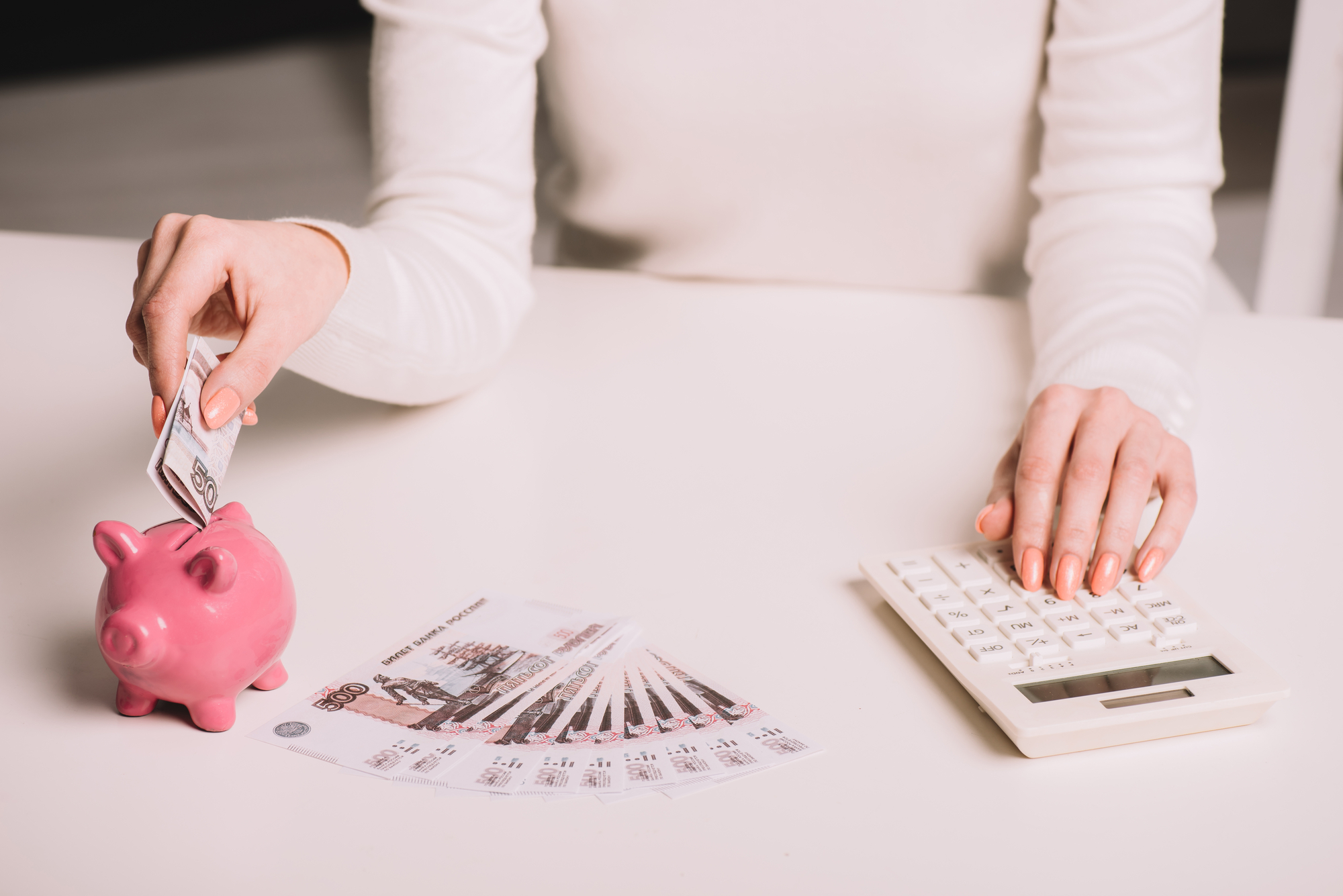 woman at white desk putting money notes into a pink piggy bank with one hand and using calculator with the other with a fan of money notes in front of her.