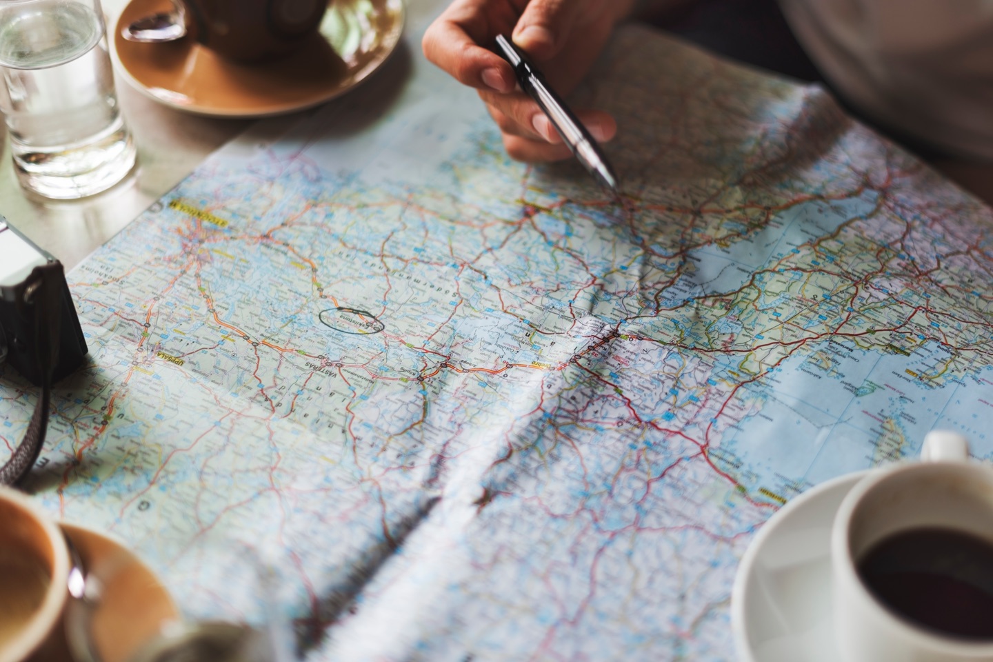 Photo. Paper map spread out. Beverages in cups and glasses around edge. Hand holding pen perched over map at top center.