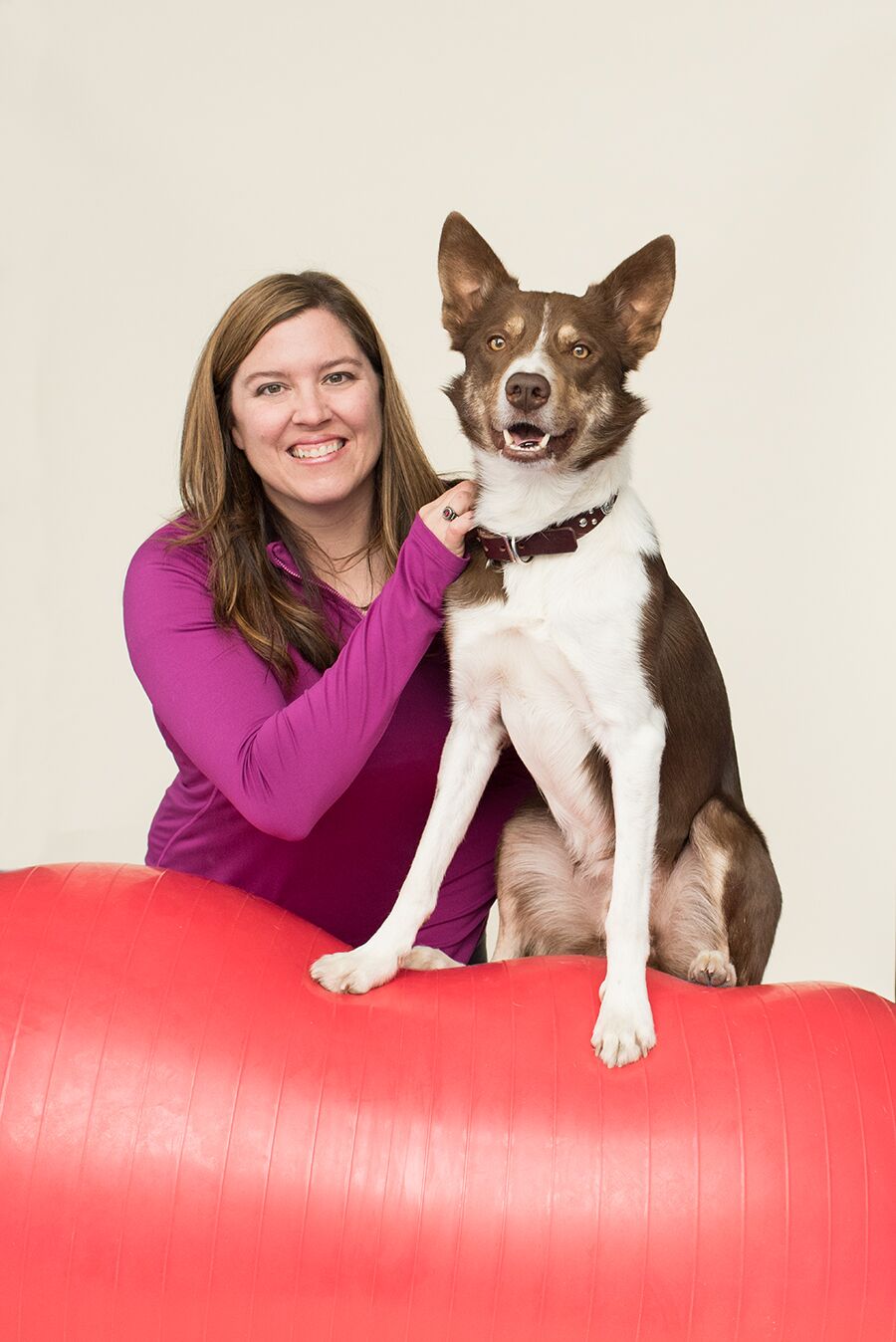 A white passing woman with long golden highlights in brown hair holds the collar of a dog sitting to the right of her. She is wearing a dark pink long sleeves shiny shirt. She has a ring on the ring finger of the hand holding the dogs collar. She is smiling. The dog is a red tri smooth border collie with prick ears, a white chest and belly and white front legs. He is sitting on red inflatable fitness peanut. He is smiling. Both the woman and dog are looking straight into the camera.