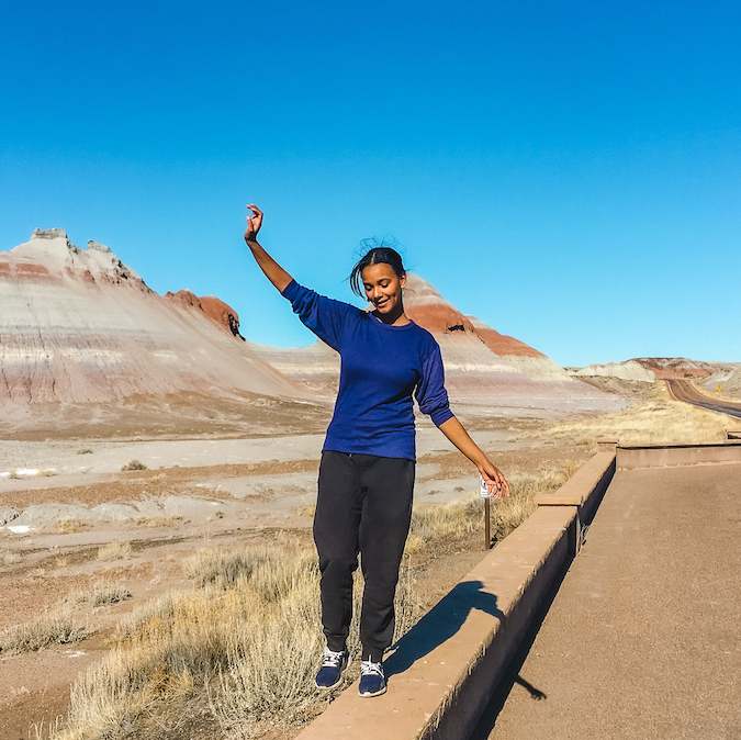 Black woman walking along the edge of a national park road, with blue mesa rock structures towering behind her
