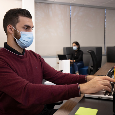 In a office workplace, a person is wearing a mask and working on a laptop computer. There is another person in the background doing the same.