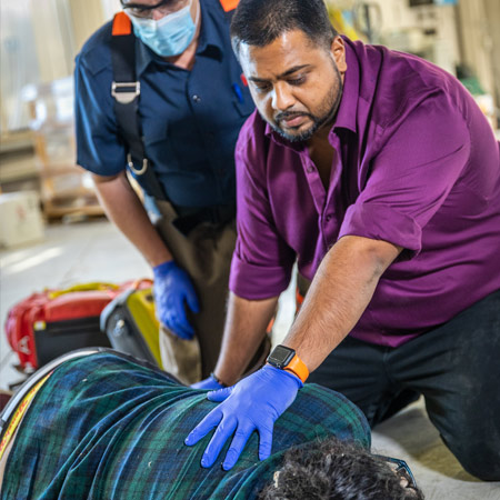 A person in a purple shirt with a worried and scared face accompanying a person being transferred to an ambulance because of possible opioid poisoning. An emergency staff behind the person is wearing a mask and gloves. 