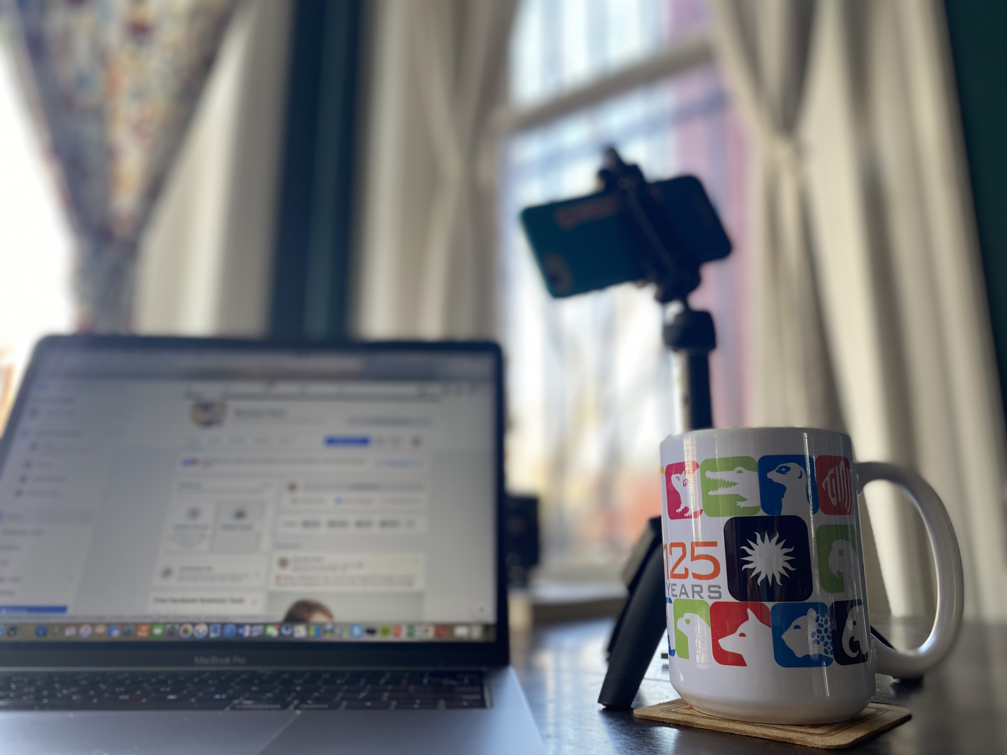 A laptop, tripod with smart phone mounted and coffee cup are pictured before a background of creamy white curtains.