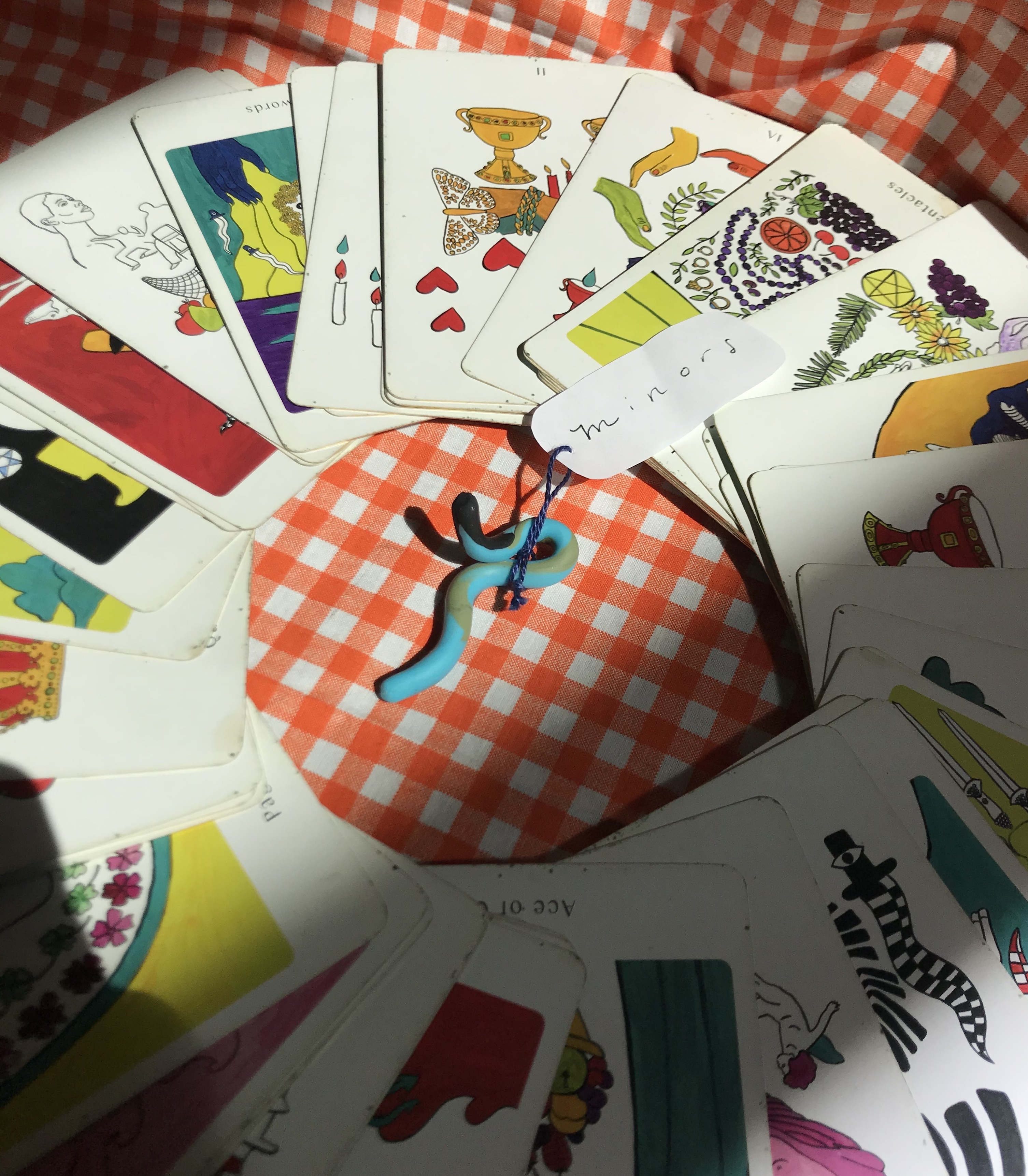 a circle of minor arcana cards, face up, surrounds a clay squiggle with a tag on it that reads: Minors. beneath the cards is a background of orange gingham