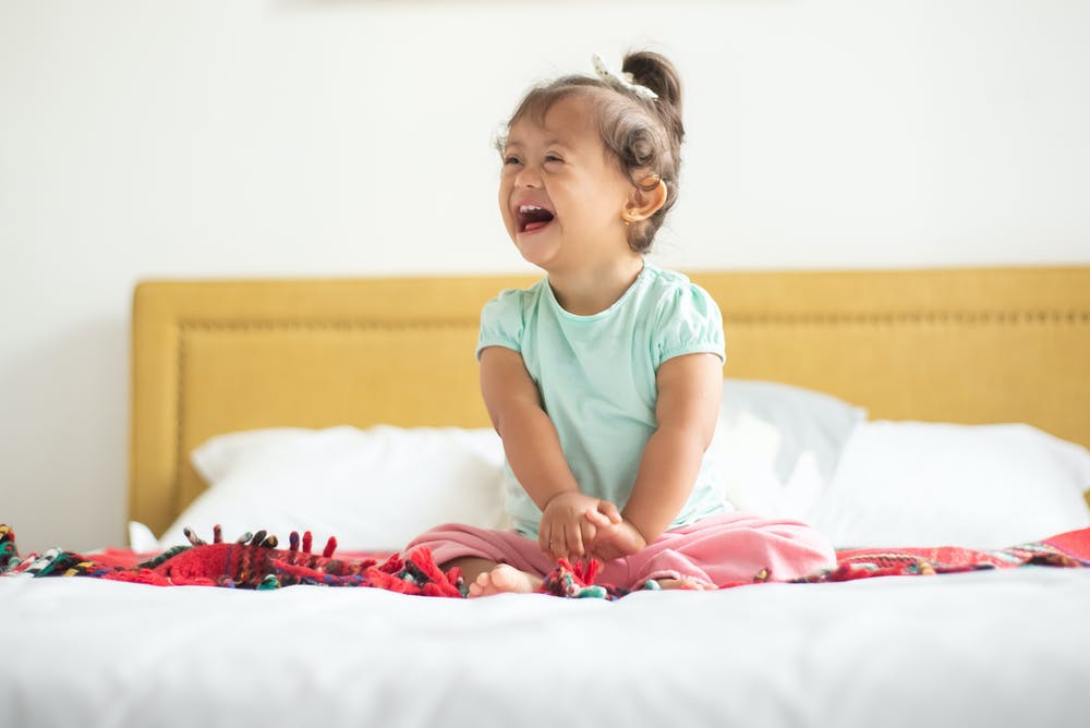Photo of 2 year old girl with down syndrome, wearing a light green shirt and pink pants, sitting on a bed. The child is laughing and smiling up to the left.