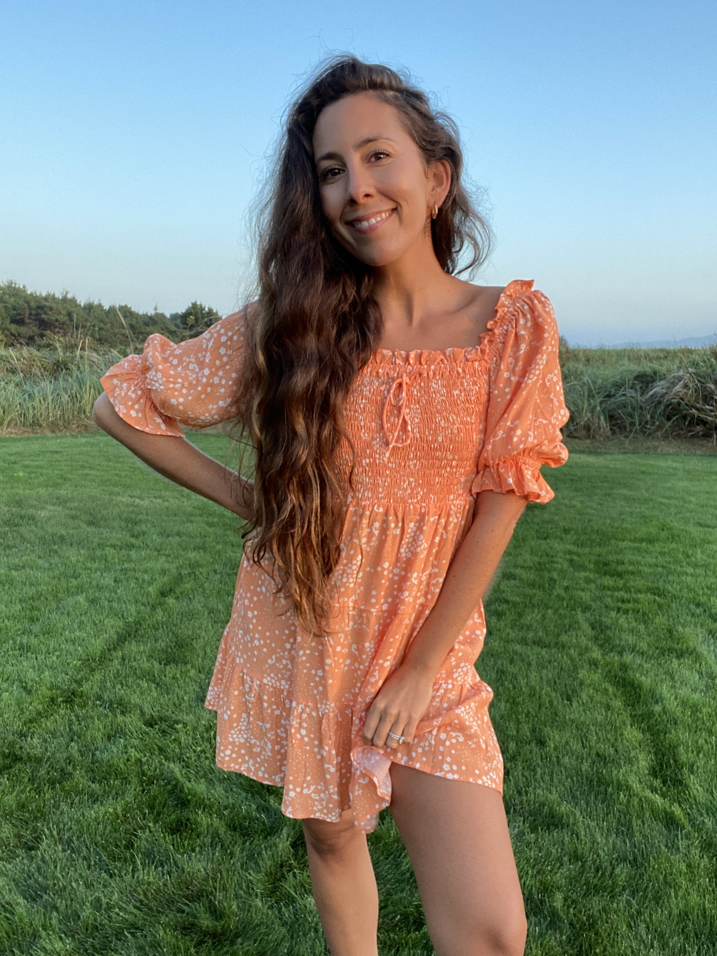 A woman standing in a field of grass feeling blissfully happy and in her worth. Wearing a orange daisy dress this is sustainably made.