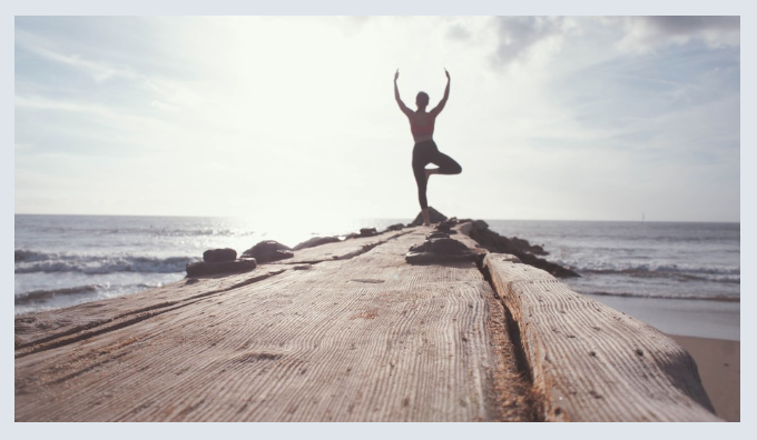 Person in yoga tree pose at the end of a pier, looking at beach.