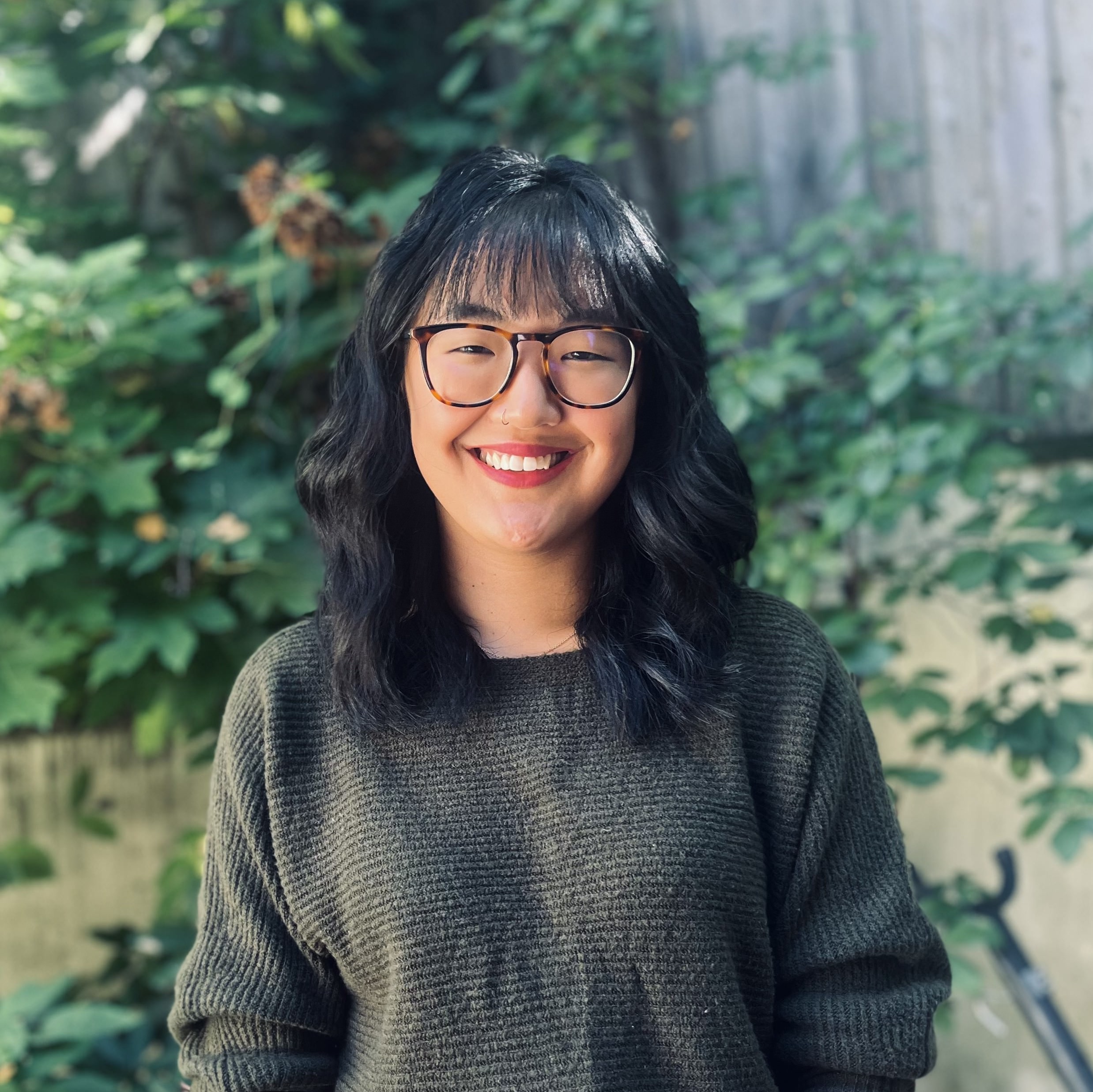 Agent and instructor Amy Bishop-Wycisk stands before a plant filled background, wearing tortoise shell glasses and smiling with shoulder length black hair.