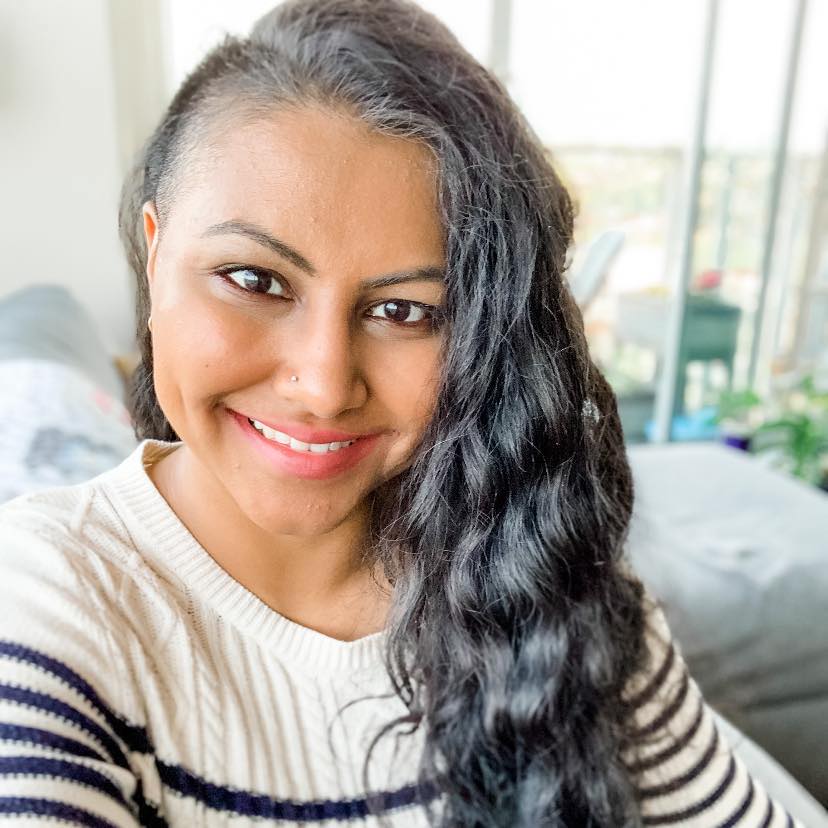 Sareeta smiles at the camera, seated on a grey couch, her long wavy hair falling past her left shoulder. She is wearing a knit white sweater with blue horitizontal stripes across the sleeves and torso. 