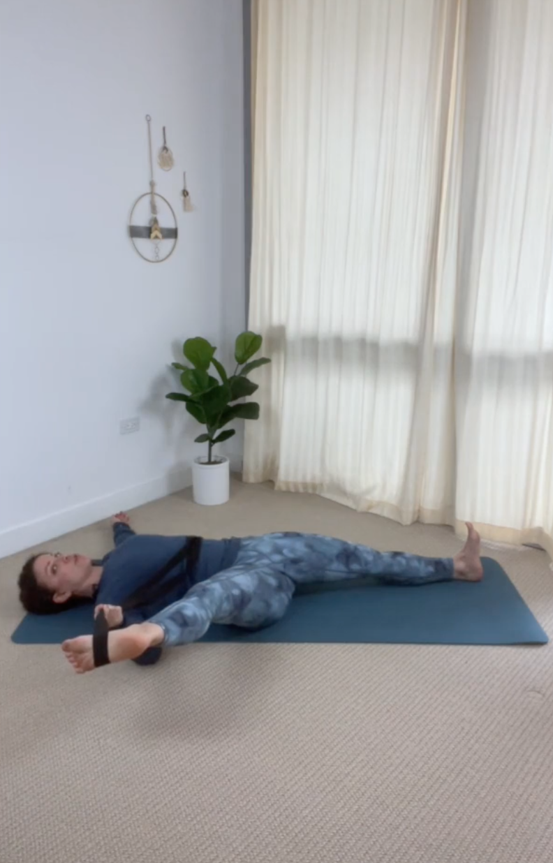 Teacher reclined with right leg outstretched to the side with yoga strap around her foot