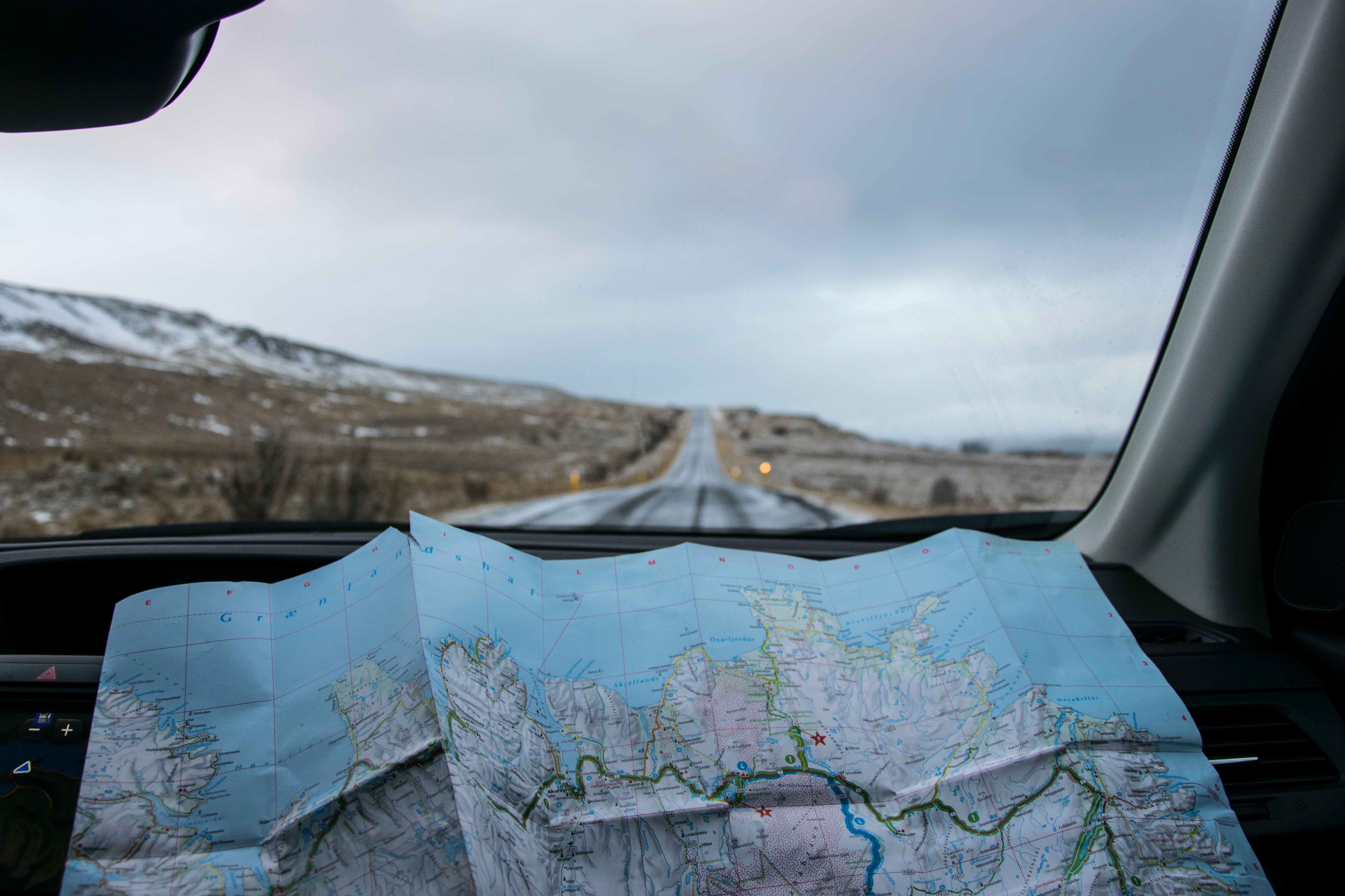 Car dashboard with a map spread out in the foreground and a road stretched snow capped mountain in the background