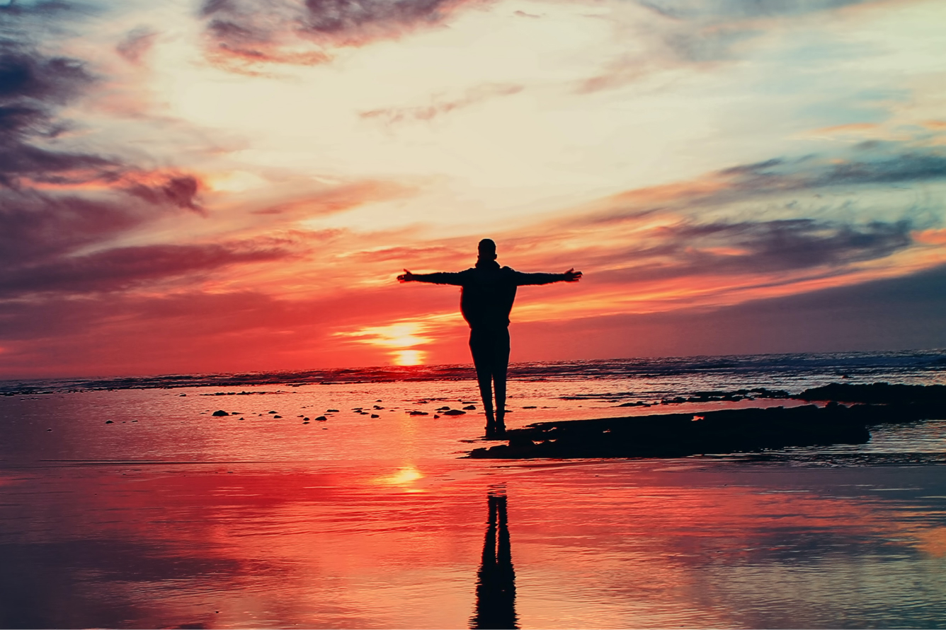 A person stands on the sand of a beach at sunrise with their arms stretched out