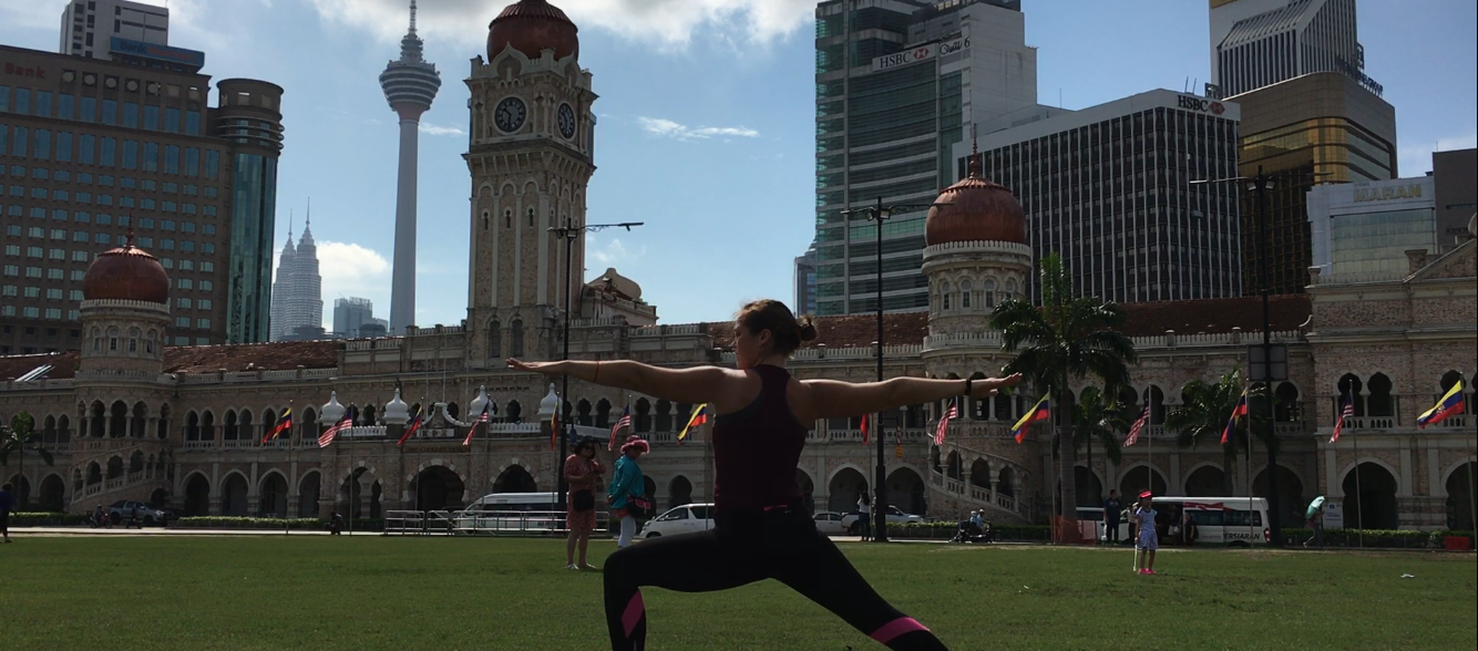 Woman stands in warrior 2 yoga pose with arms outstretched in front of Kuala Lumpur skyline.