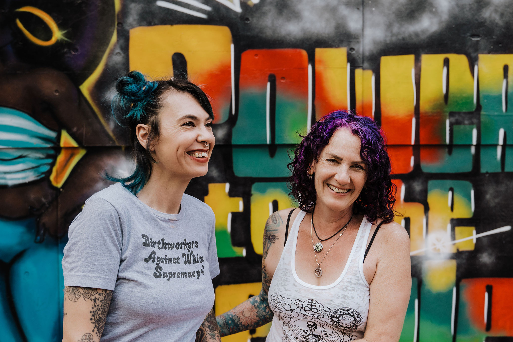 Nickie and Juli Tilsner stand in front of a colorful mural that says &quot;Power to the people&quot;. They are smiling, Juli&#39;s hand is on Nickie&#39;s arm, and Nickie is looking to the right.