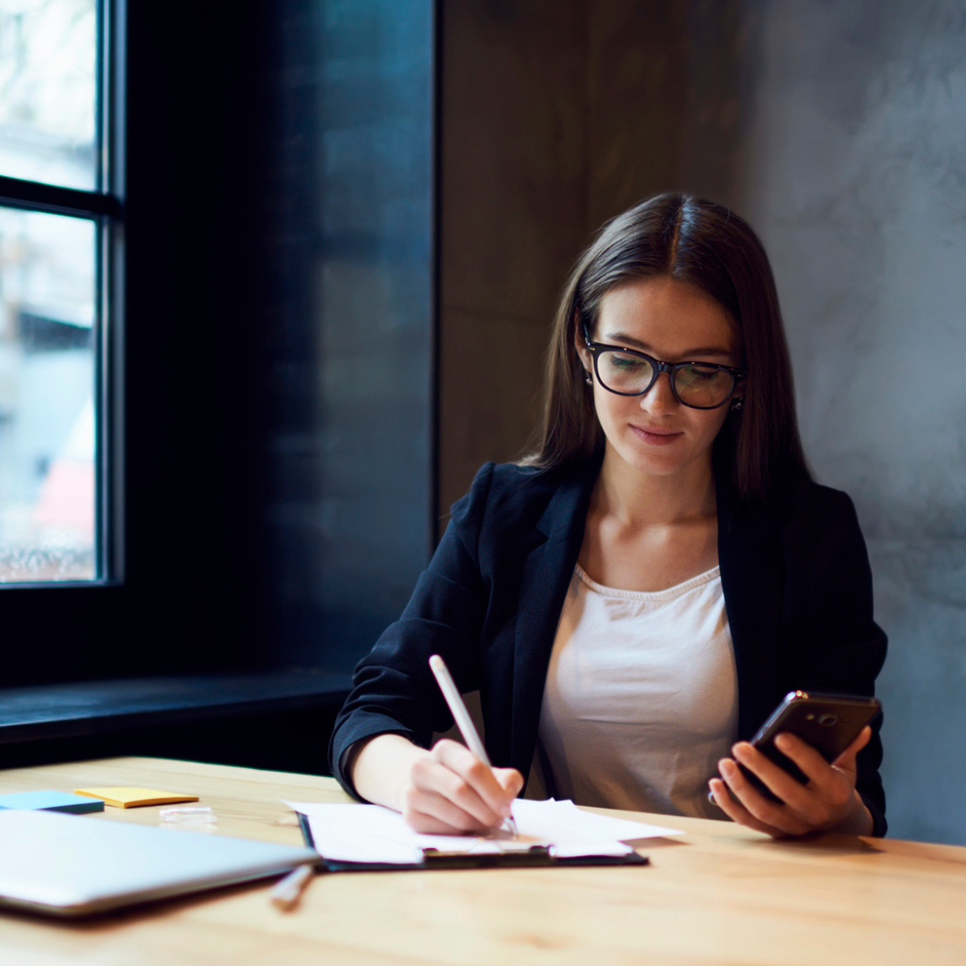 A woman with glasses, wearing a dark blue business jacket and white shirt, sits at a table with a note pad and her phone, making notes.