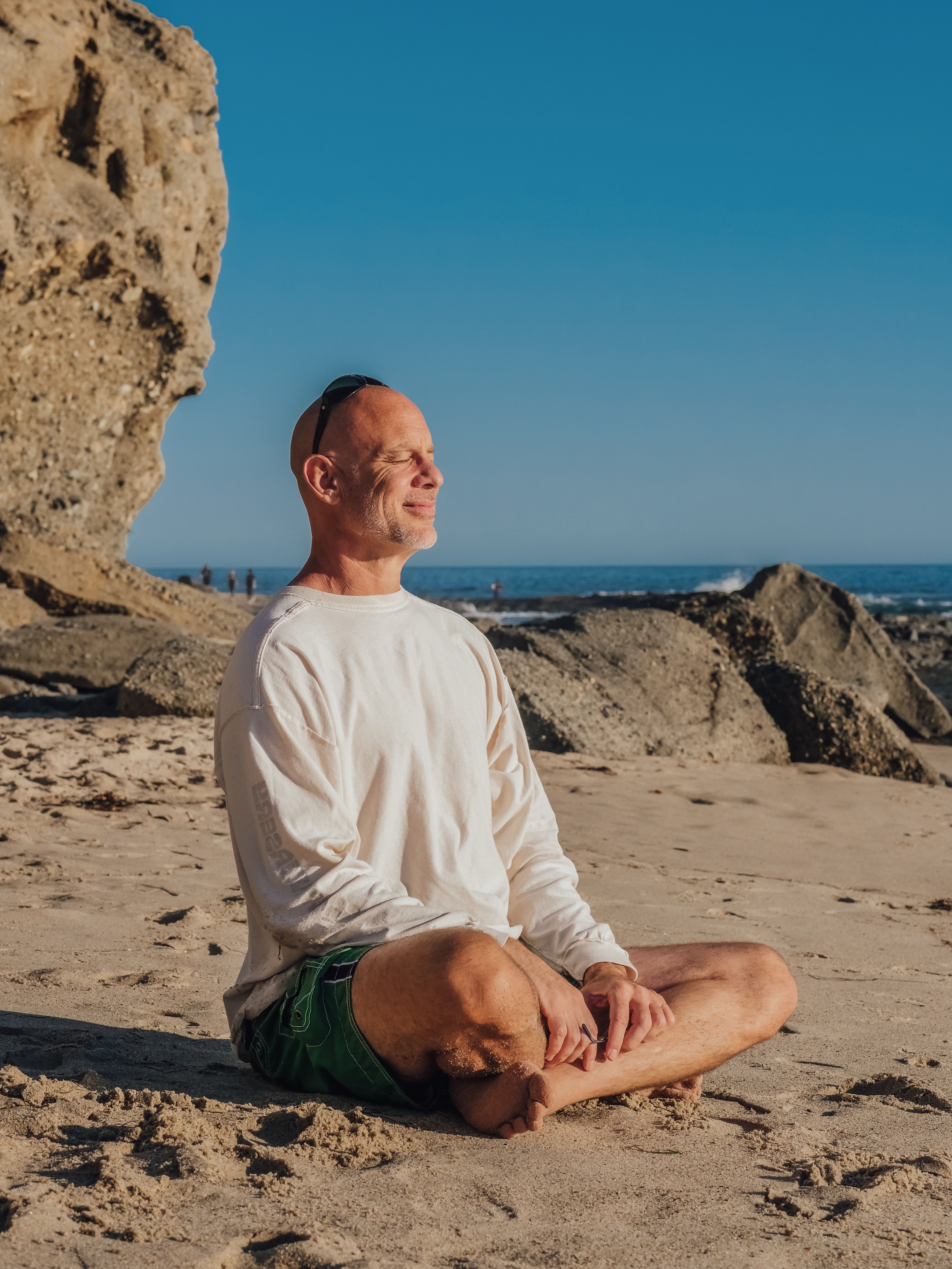 middle aged man sitting in meditation on beach in long sleeved tee shirt and shorts