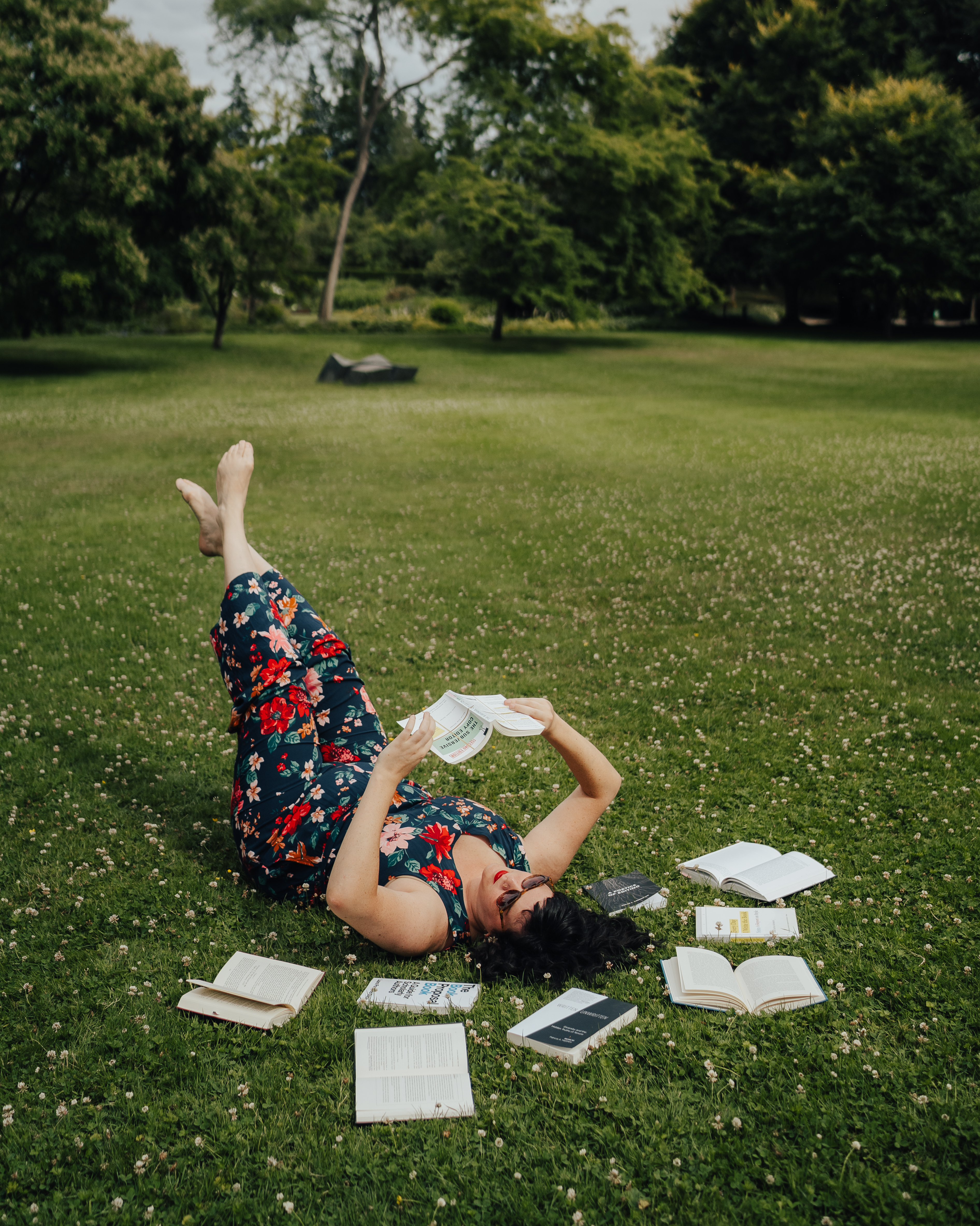 A picture of Letitia Henville, lying on the grass, reading a book. She is wearing sunglasses and a colourful floral jumpsuit.