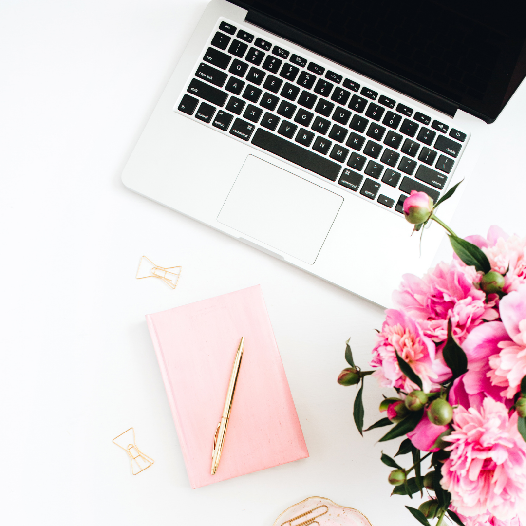Picture Of laptop, pink journal with a gold pen on top, and pink flowers on a white desk