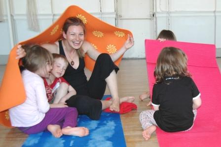 A yoga teacher holds a yoga mat over two children as they smile and laugh.