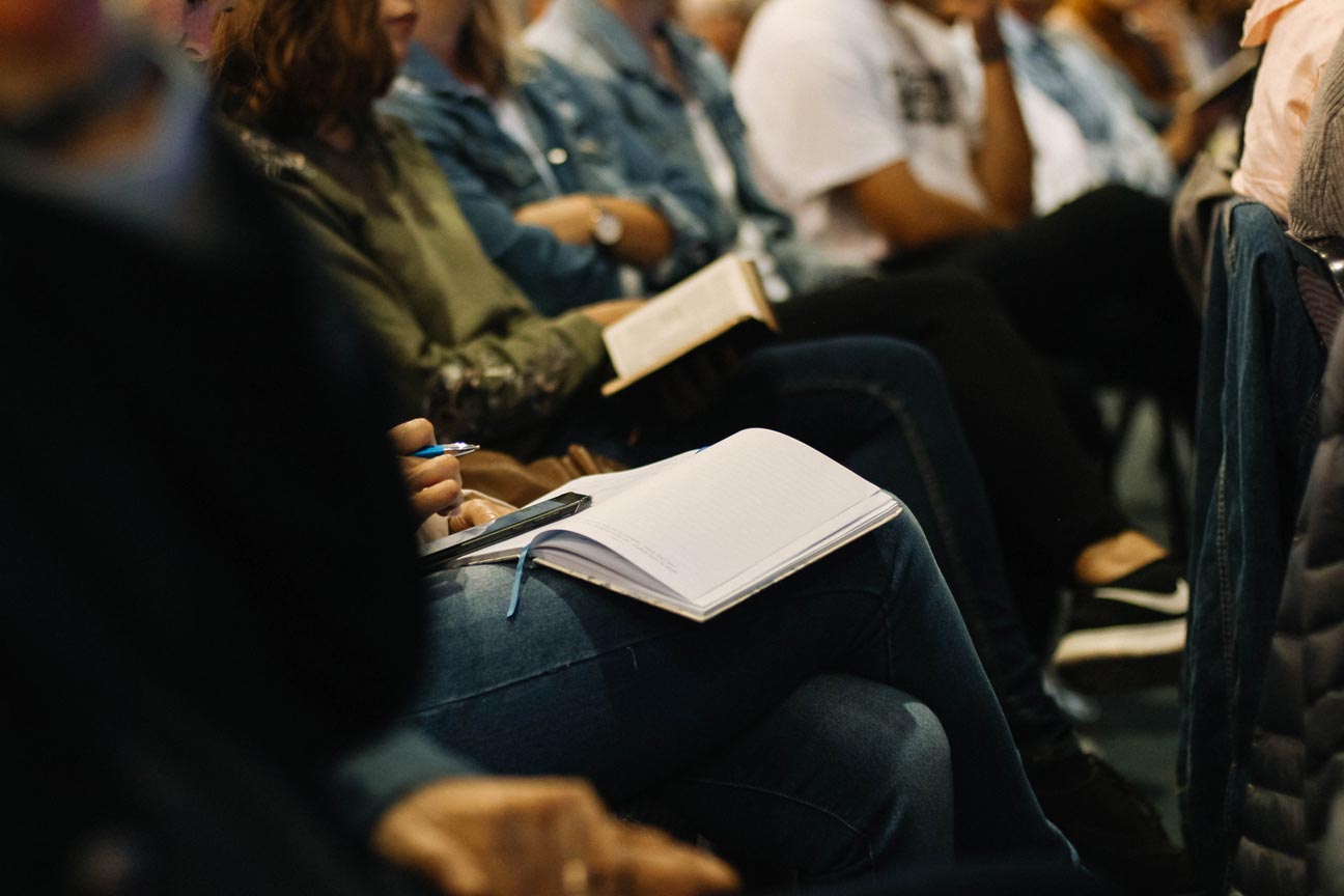 A group of people sit listening to a lecture. A few of the audience members are taking notes in notebooks.