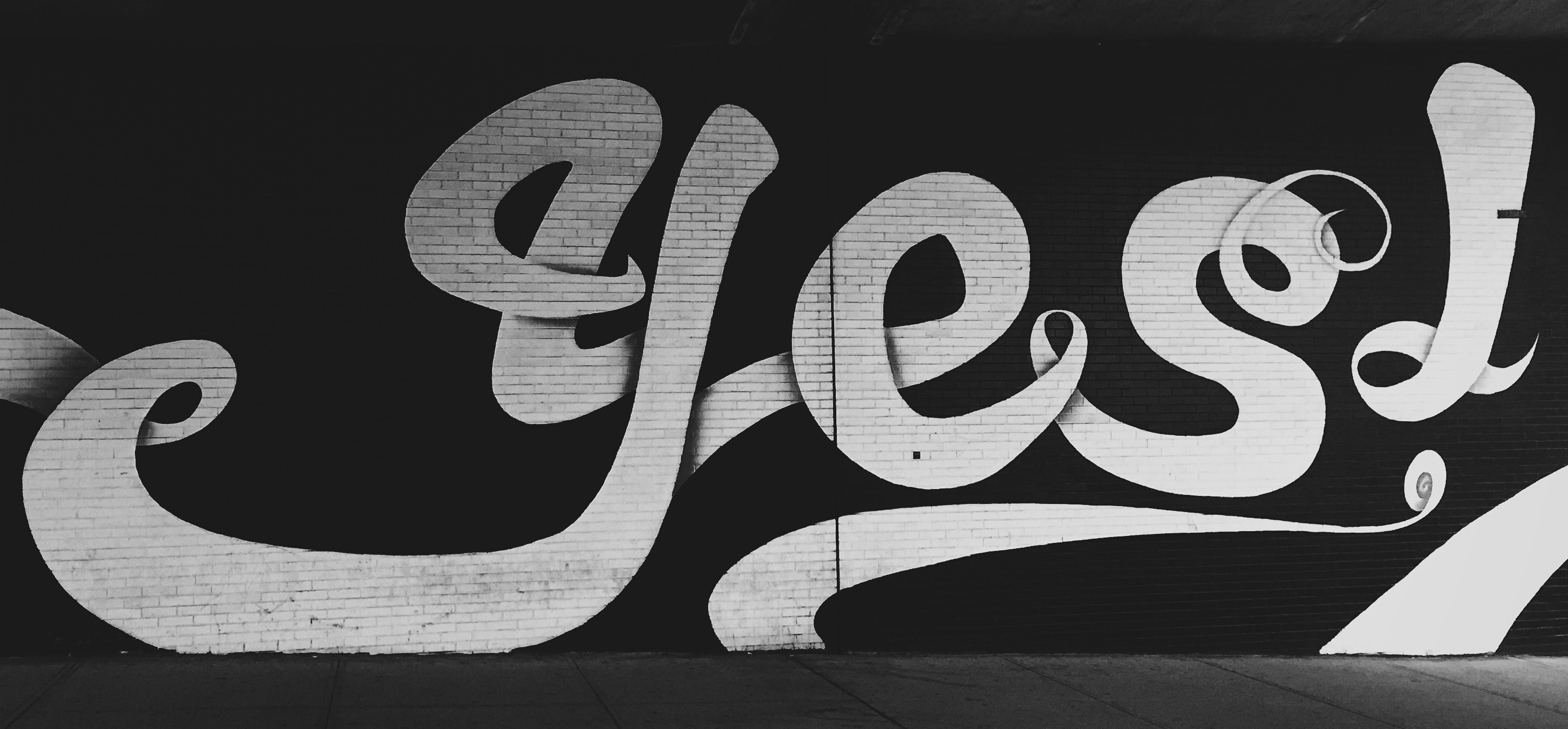 "yes!" painted in white swirly text on a black brick wall