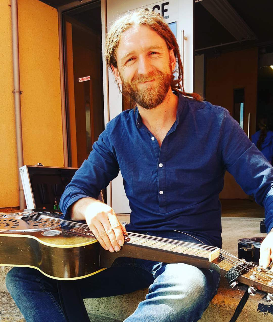 Phillip Henry sits holding a Dobro slide guitar, smiling