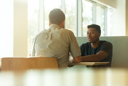 A teacher and a student standing at a whiteboard at the front of a classroom, presenting to a group of students seated at tables.