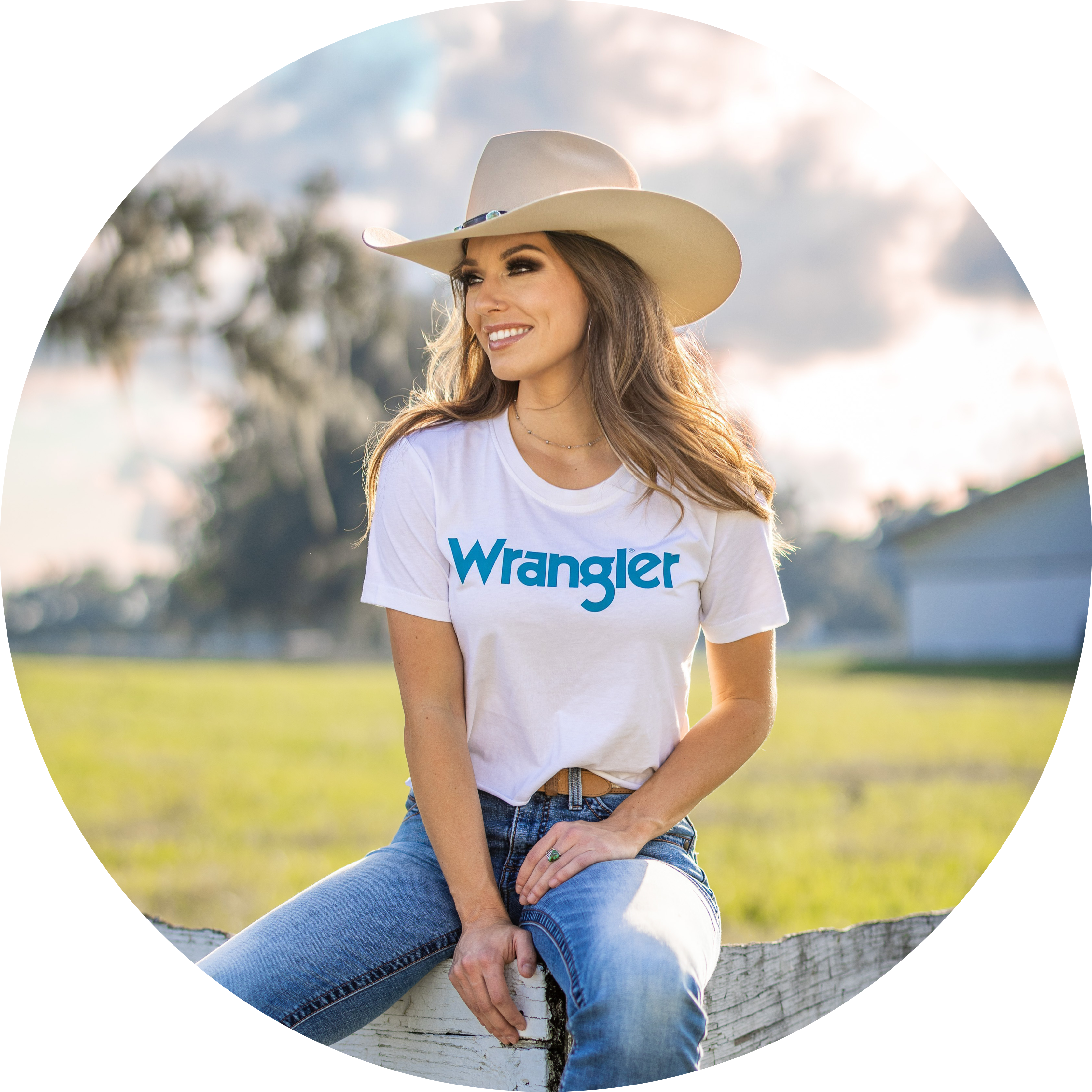 Olana sitting on a wooden fence looking to the left, wearing a cowgirl hat, white Wrangler shirt, and blue jeans