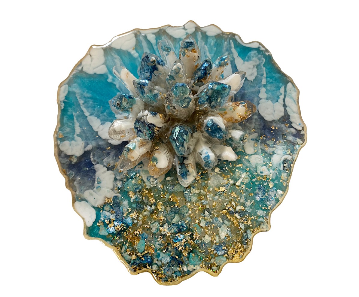 A standalone resin art piece is showcased, reminiscent of a blooming flower or a coral reef from an enchanting underwater world. It features a dynamic blend of blues and whites with gold accents, complemented by a constellation of what appears to be glittering stones or glass in the center. This handcrafted piece radiates a serene yet luxurious aesthetic, proudly bearing the signature 