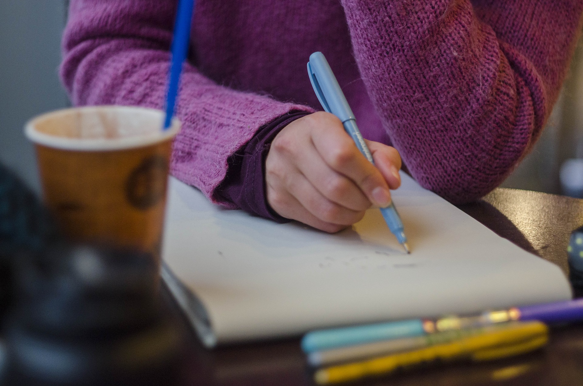 Image of a hand writing with a blue pen on a notepad, with a coffee cup and pens on the desk around