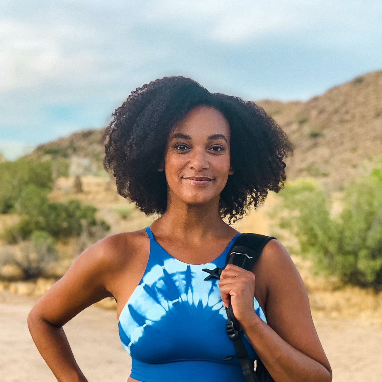 Black Hiker getting outdoors in Joshua Tree National Park holding a backpack, hand on her hip, wearing an afro